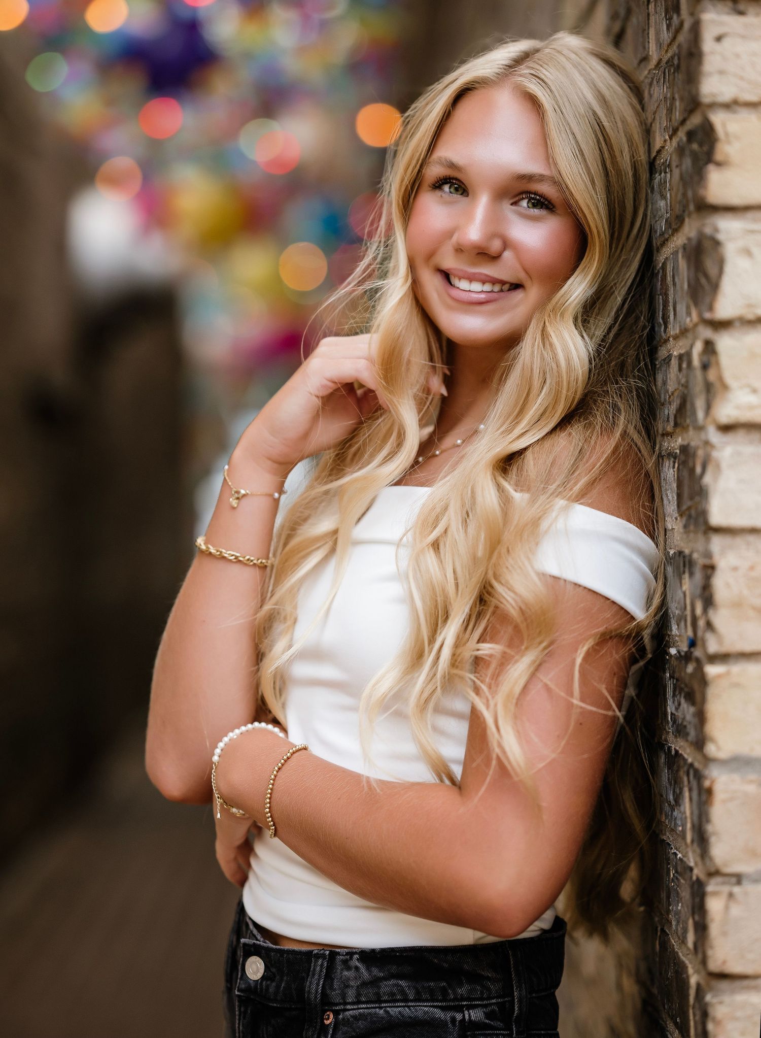 Portrait of a person in a white off-shoulder top and dark jeans leaning against a brick wall with bokeh lights in background.
