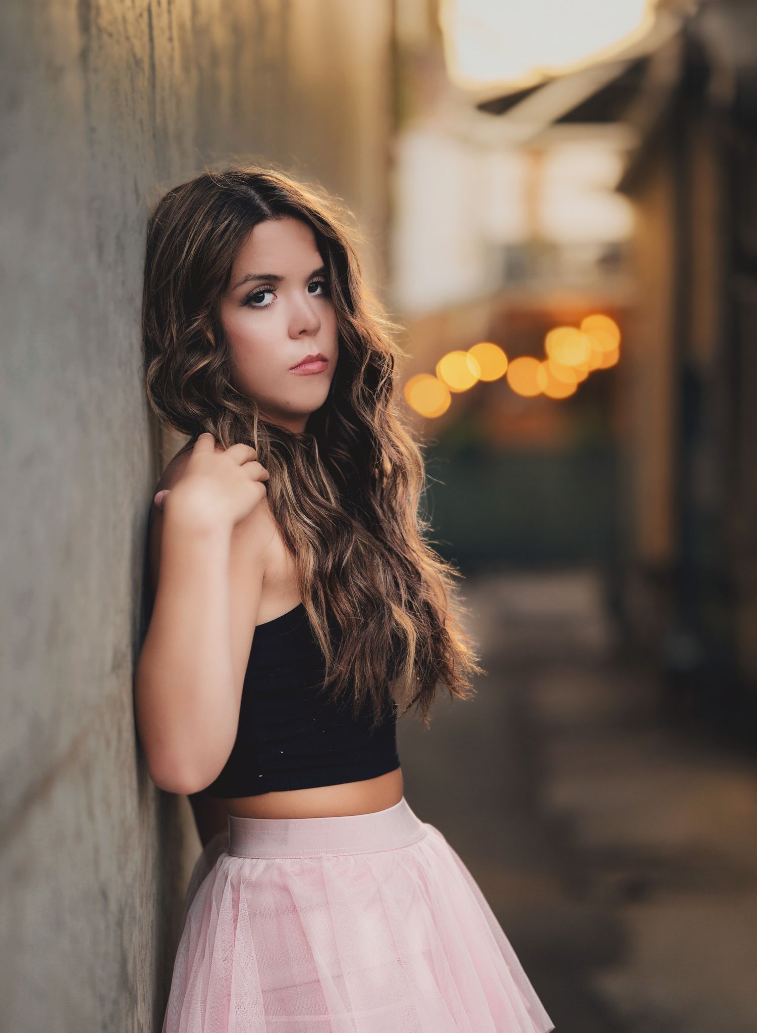A person in a black crop top and pink tulle skirt poses against a wall with blurred bokeh lights in the background.