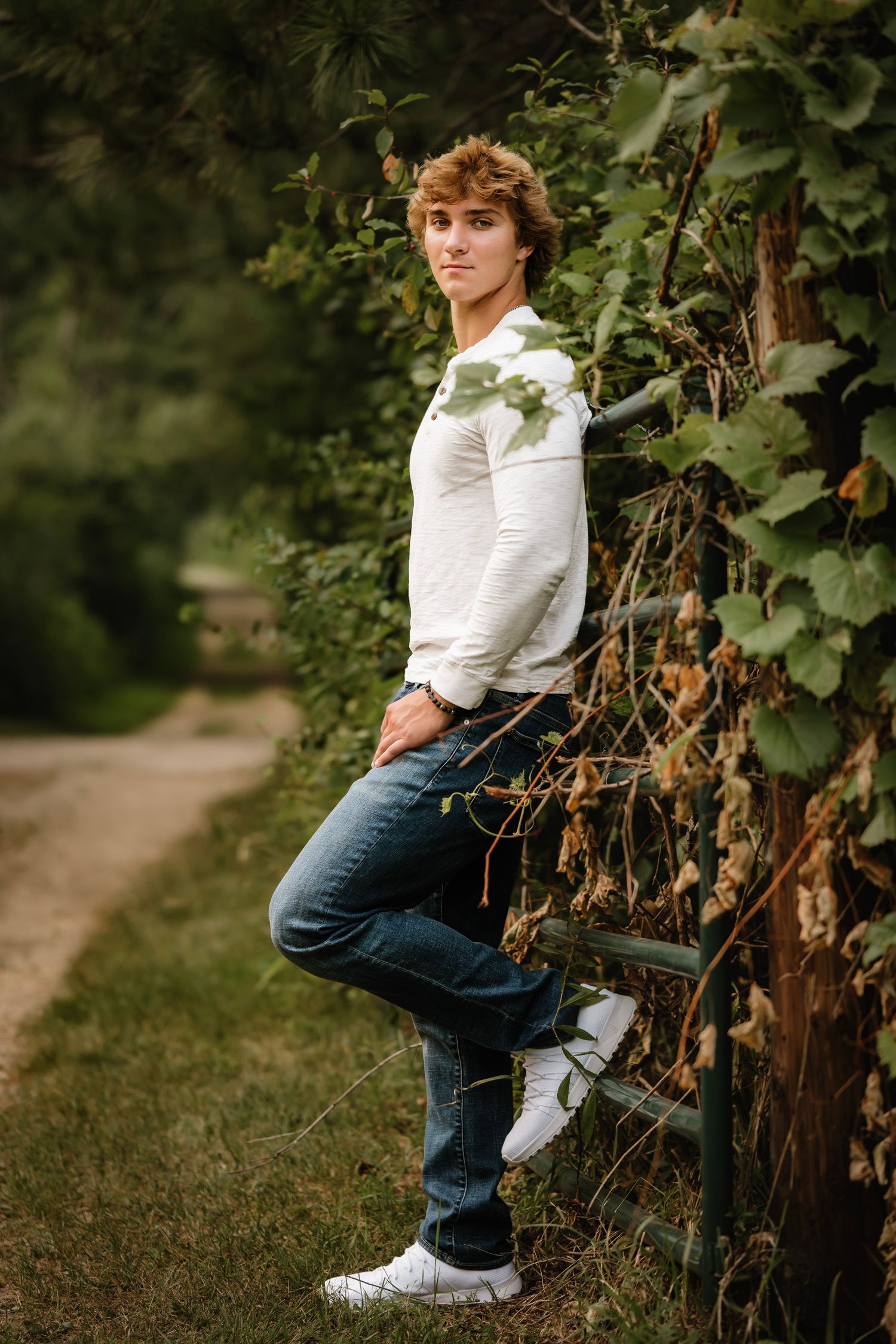 Young man in white shirt and jeans leaning against a vine-covered fence on a scenic outdoor path.