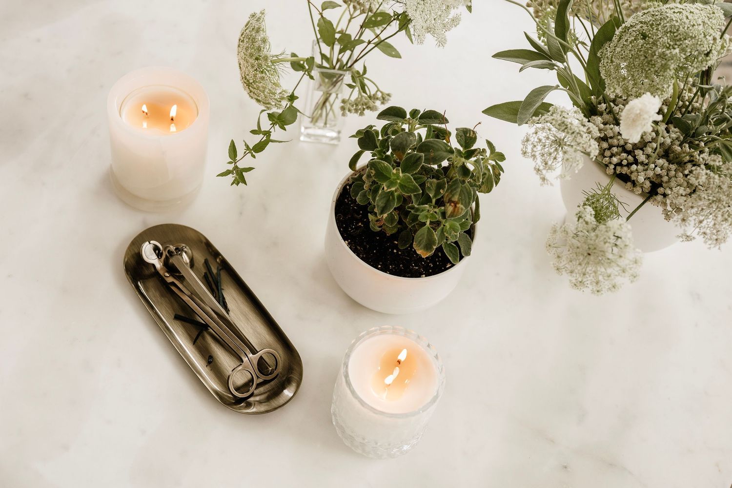 Elegant flat lay with lit candles, potted plant, greenery, and silver spoons on a white marble surface.