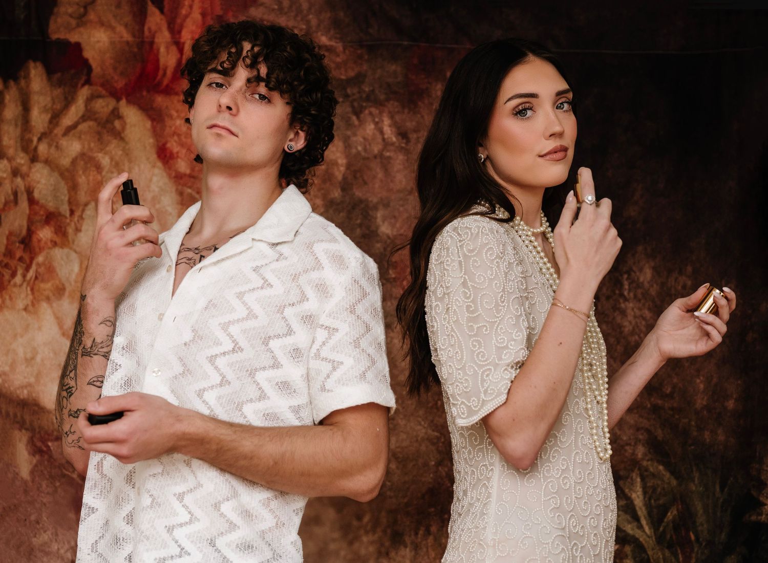 A young man and woman pose back-to-back holding perfume bottles, dressed in elegant white outfits against a floral backdrop.