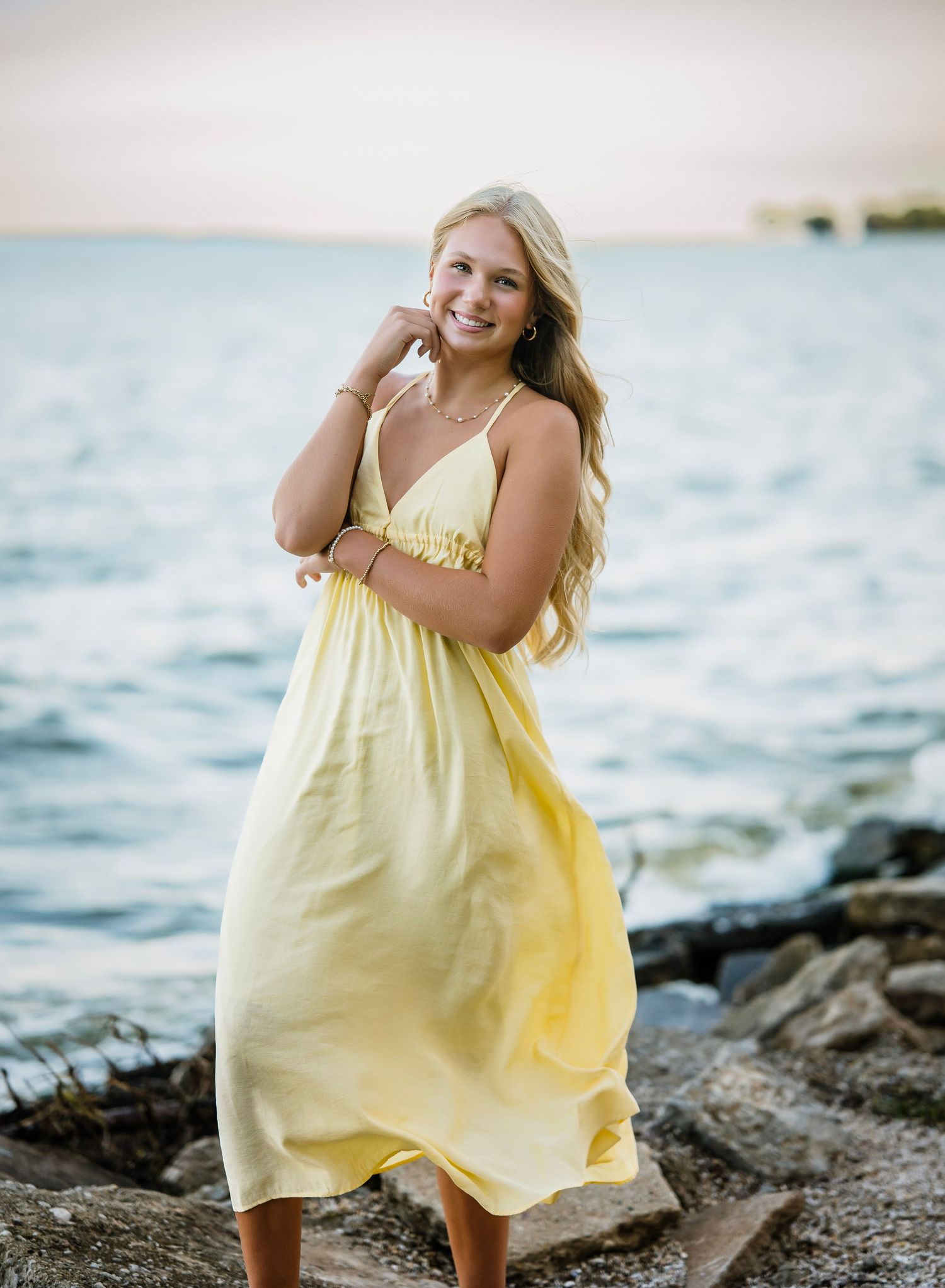 A person in a flowing yellow sundress poses playfully on rocky coastal shores at sunset.