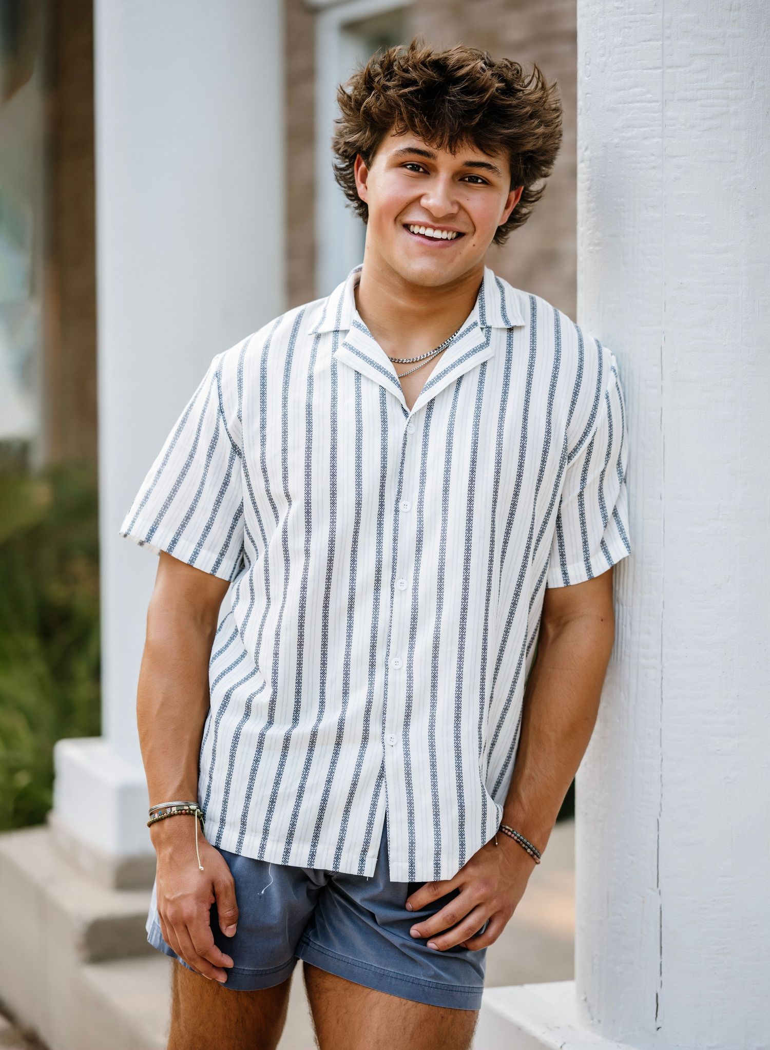 Young person in striped button-down shirt and blue shorts poses casually against white column in outdoor setting.