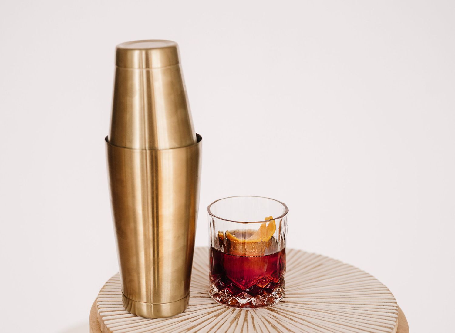 Gold cocktail shaker and red drink on a textured cream-colored surface against white background.