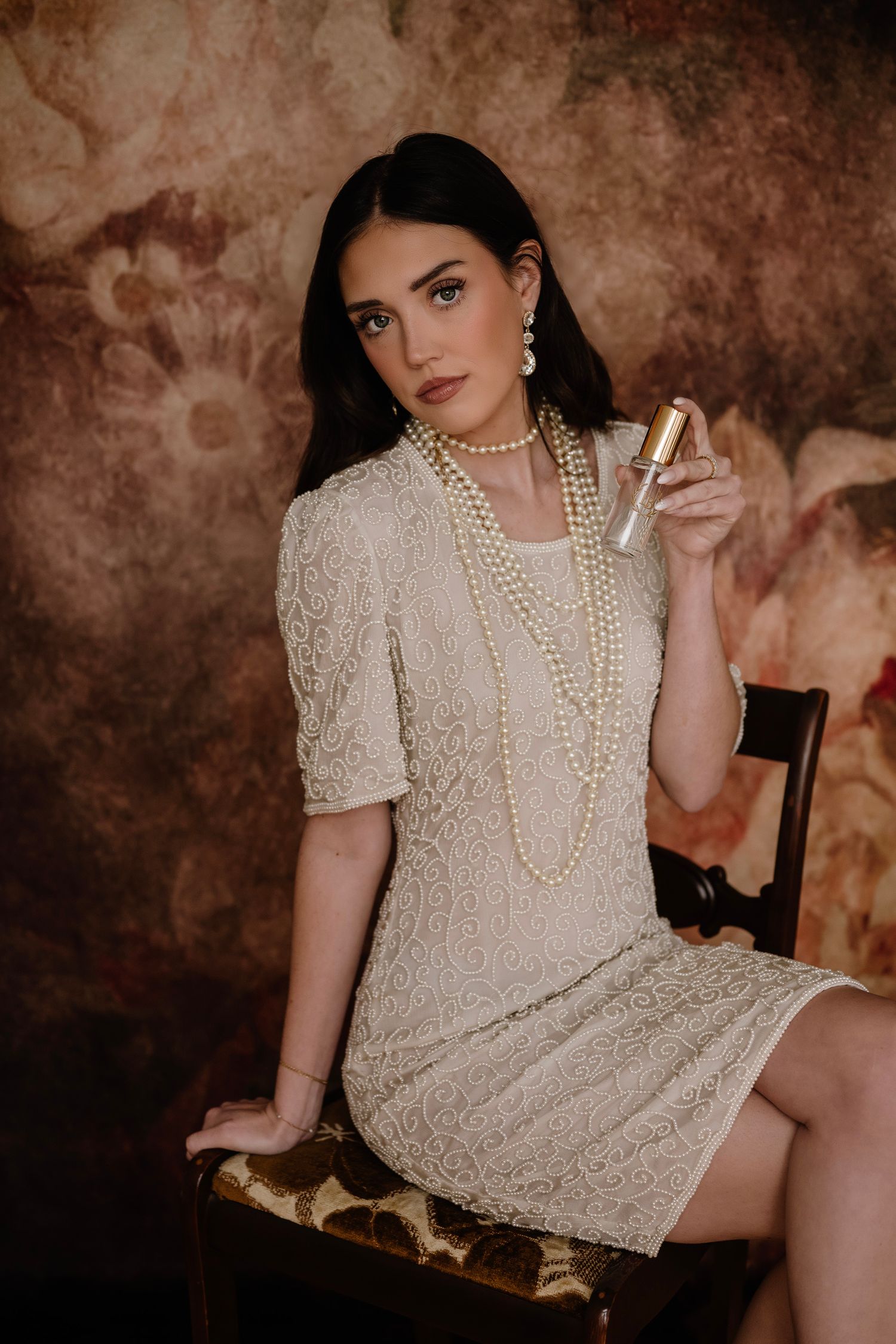 Woman in lace dress with pearl necklace holding perfume bottle, seated on chair against floral backdrop.