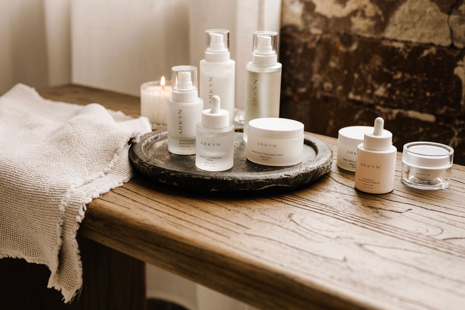 Minimalist skincare products arranged on a wooden table with lit candles and a linen towel in a cozy spa setting.