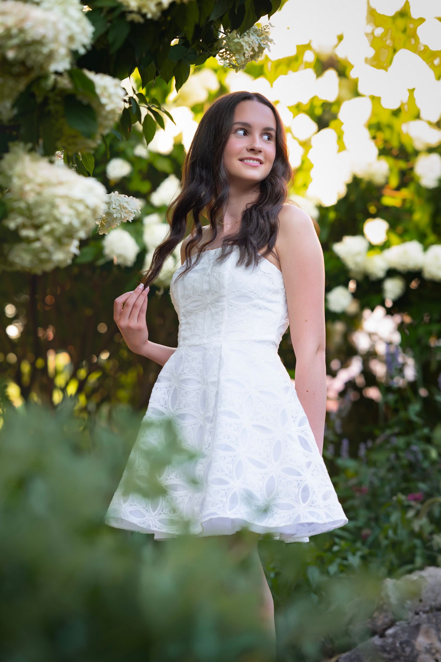 Young woman in white strapless dress smiling among blooming white hydrangea bushes in a lush garden.