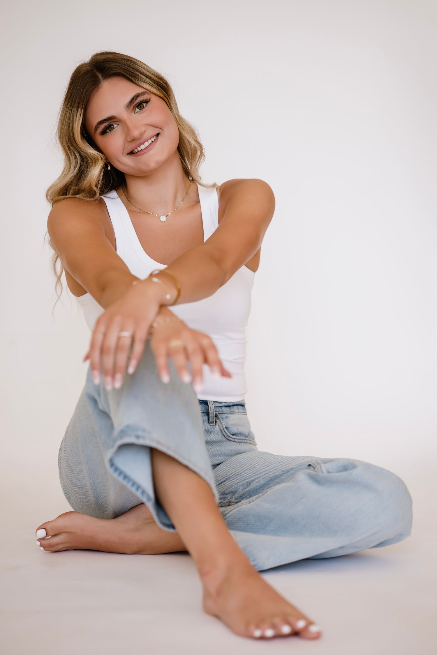 Smiling young woman in white tank top and light blue jeans sitting barefoot on white floor with arms crossed.