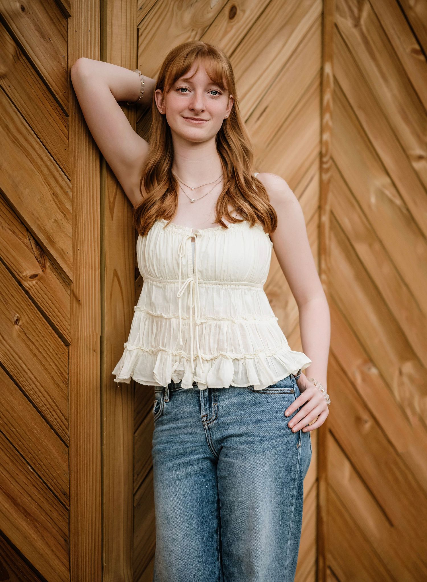 Woman in white ruffled top and jeans poses against wooden wall in natural lighting.