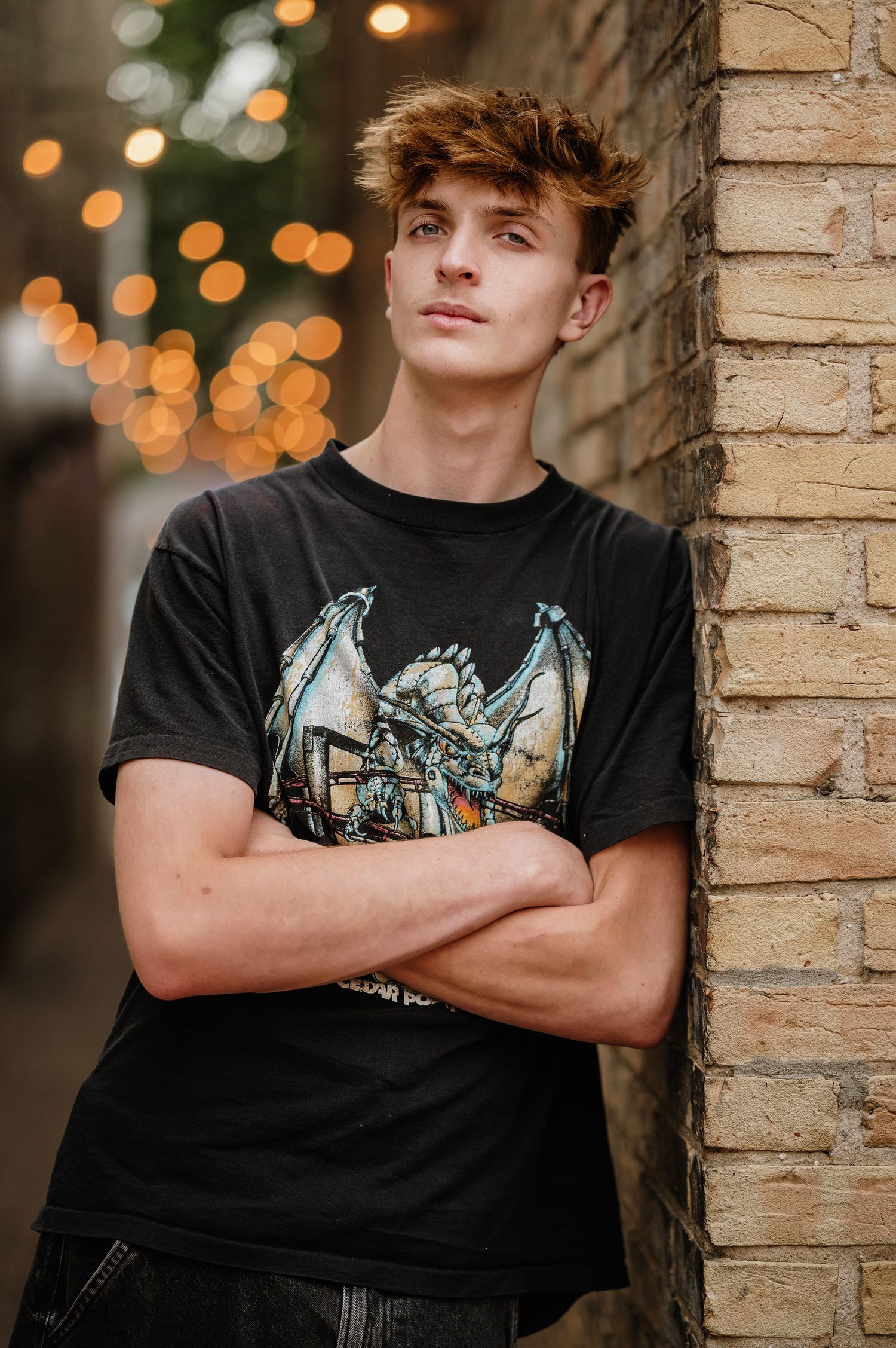 Urban senior portrait against brick wall with bokeh lights wearing graphic t-shirt with dragon design.