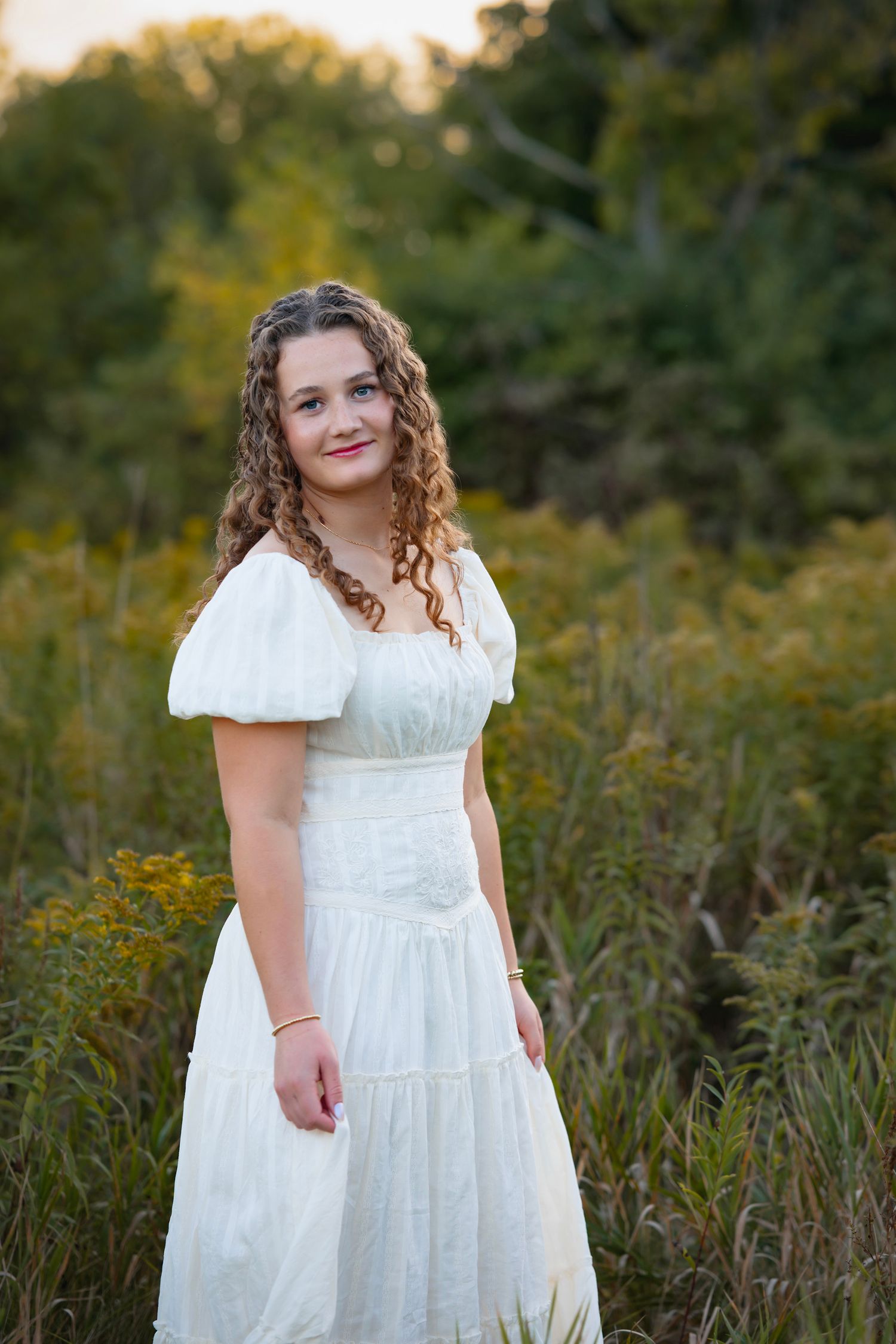 Young woman in white prairie dress standing in golden field at sunset.