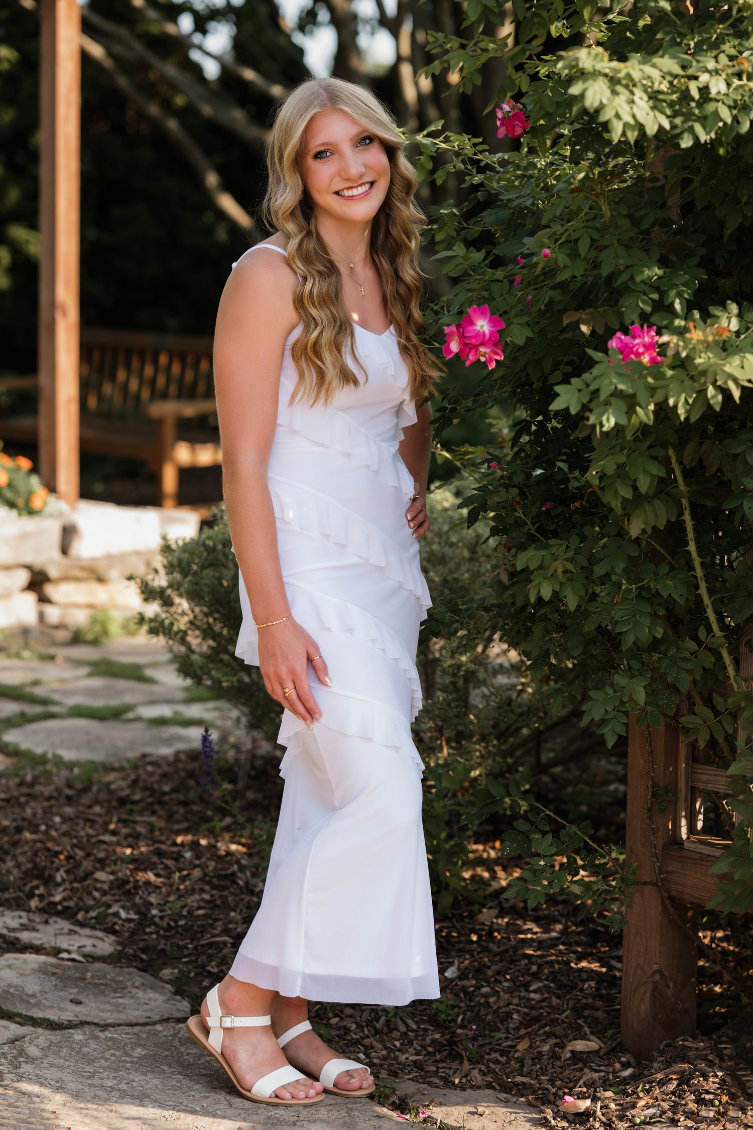 Young woman in white ruffled midi dress and sandals smiling beside pink roses in a garden.