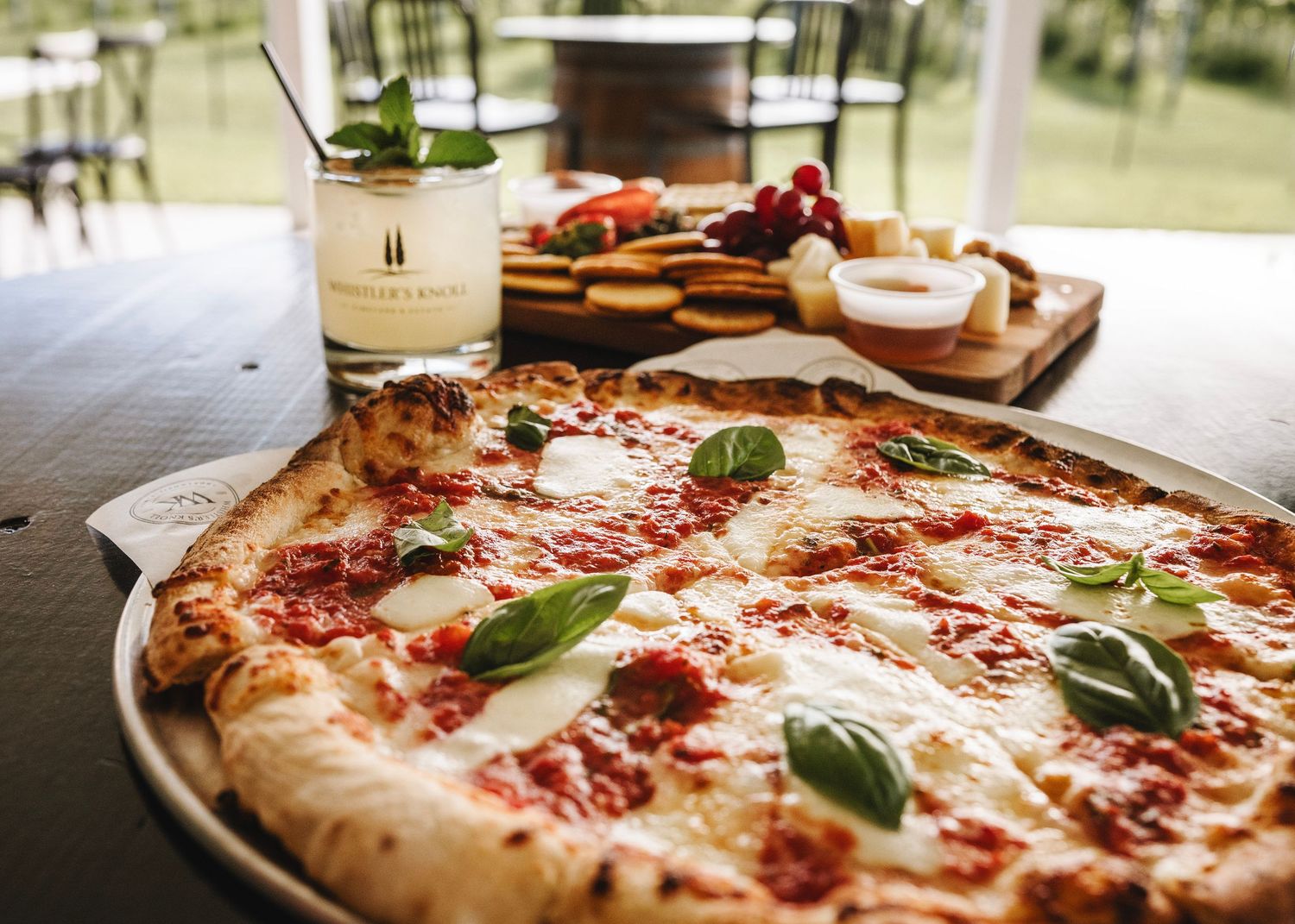 Freshly baked Margherita pizza with basil leaves on a silver tray, charcuterie board and cocktail in background.