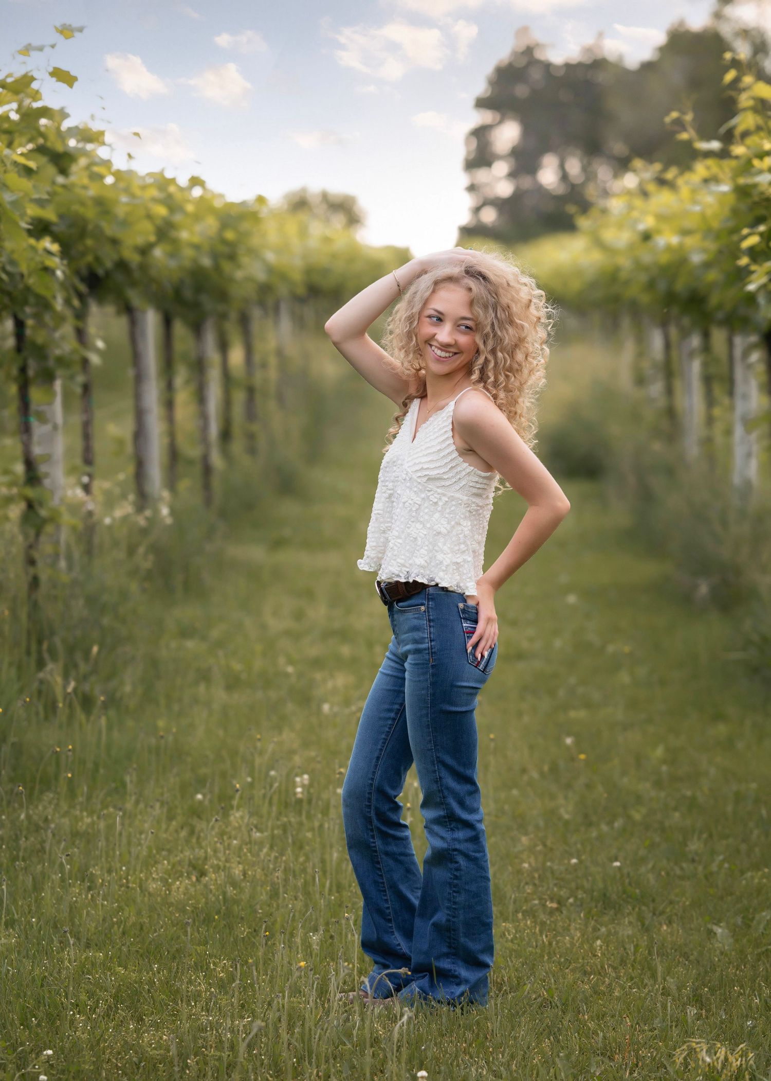 Young woman with curly blonde hair poses in blue flare jeans and white lace top between lush green vineyard rows.