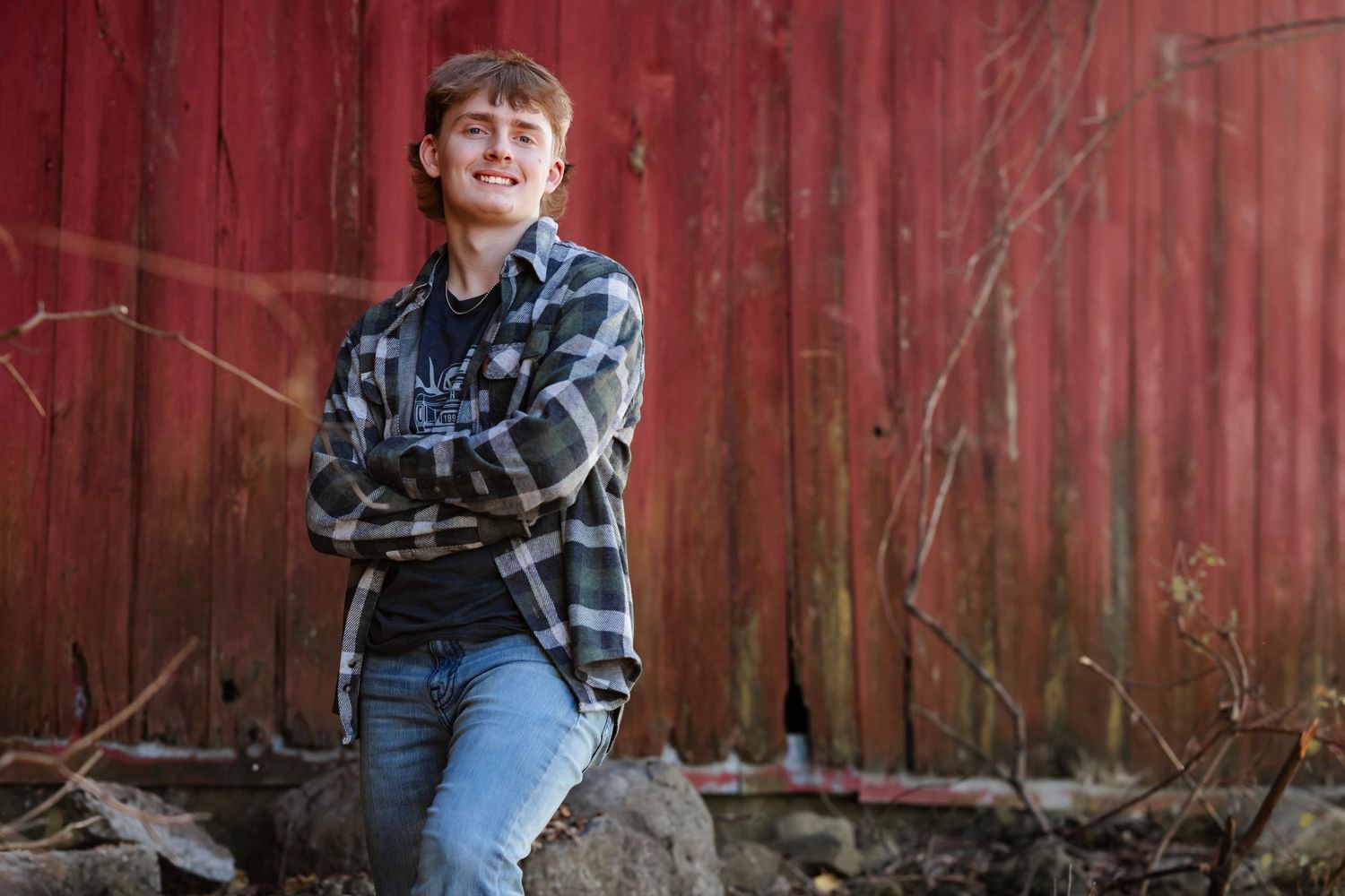 A person in a plaid flannel shirt and jeans sits casually on a rock against a rustic red barn wall.