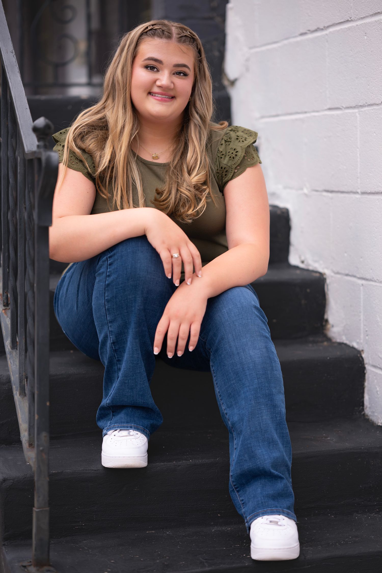Young woman with blonde hair sits smiling on black steps, wearing green top, blue jeans, and white sneakers.