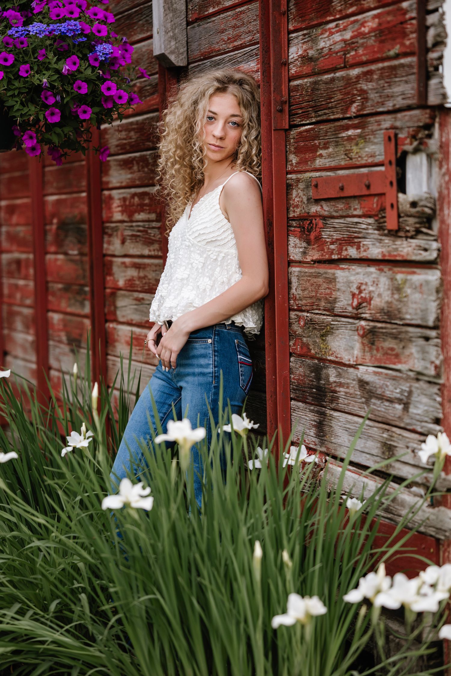 Young woman with curly blonde hair poses in blue jeans and white top against rustic red barn with white iris flowers.
