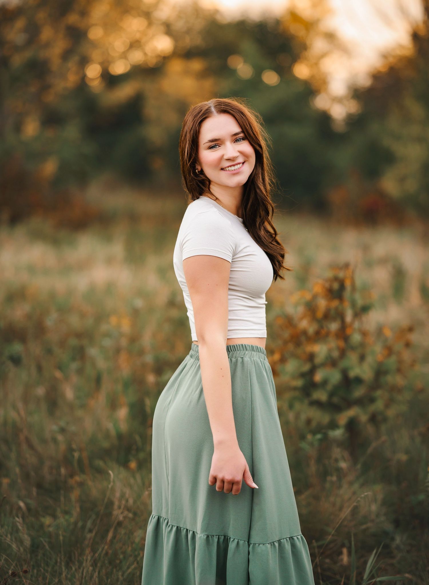 Young woman smiling in white crop top and sage green midi skirt standing in a sunlit field.
