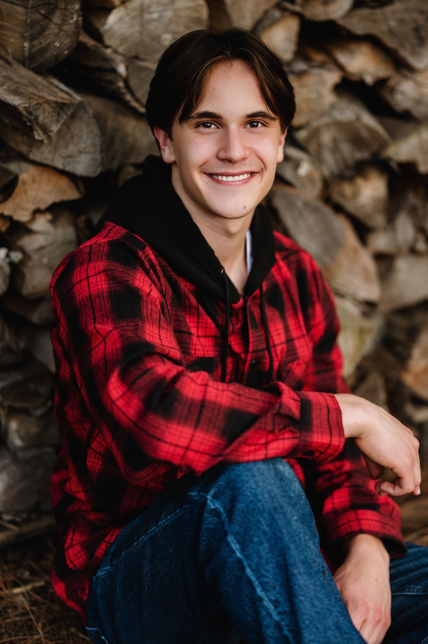 Young man in red plaid flannel shirt smiling outdoors against rustic stacked firewood background.