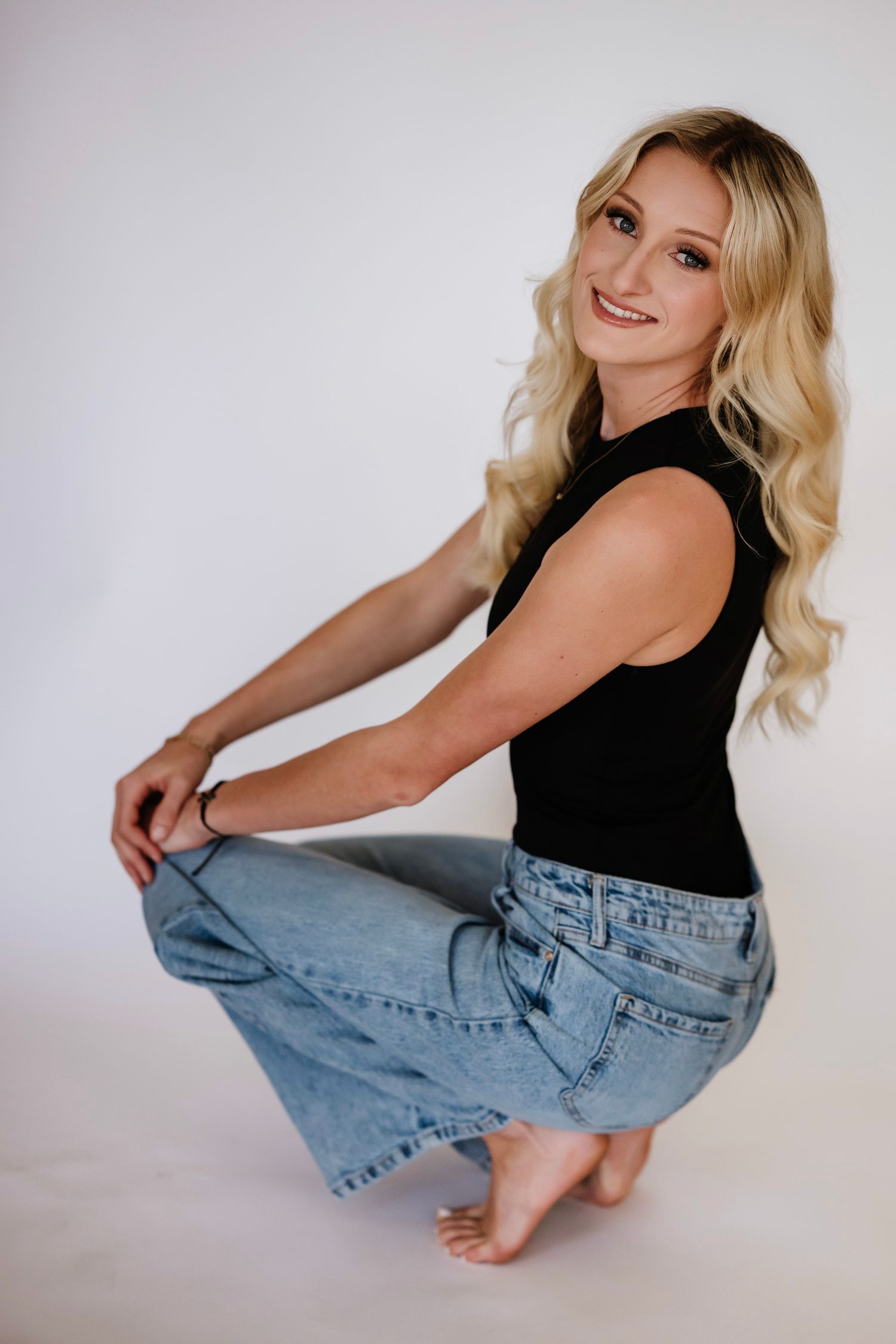 Smiling blonde woman in black tank top and oversized jeans posing barefoot on white background.