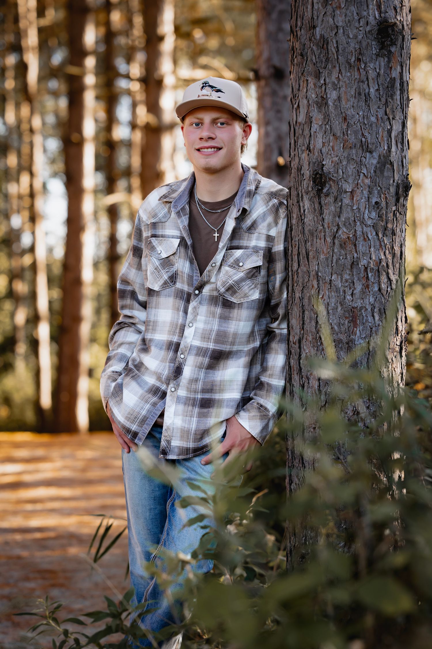 Young man in plaid flannel shirt and cap leans against pine tree in sunlit forest, smiling casually.