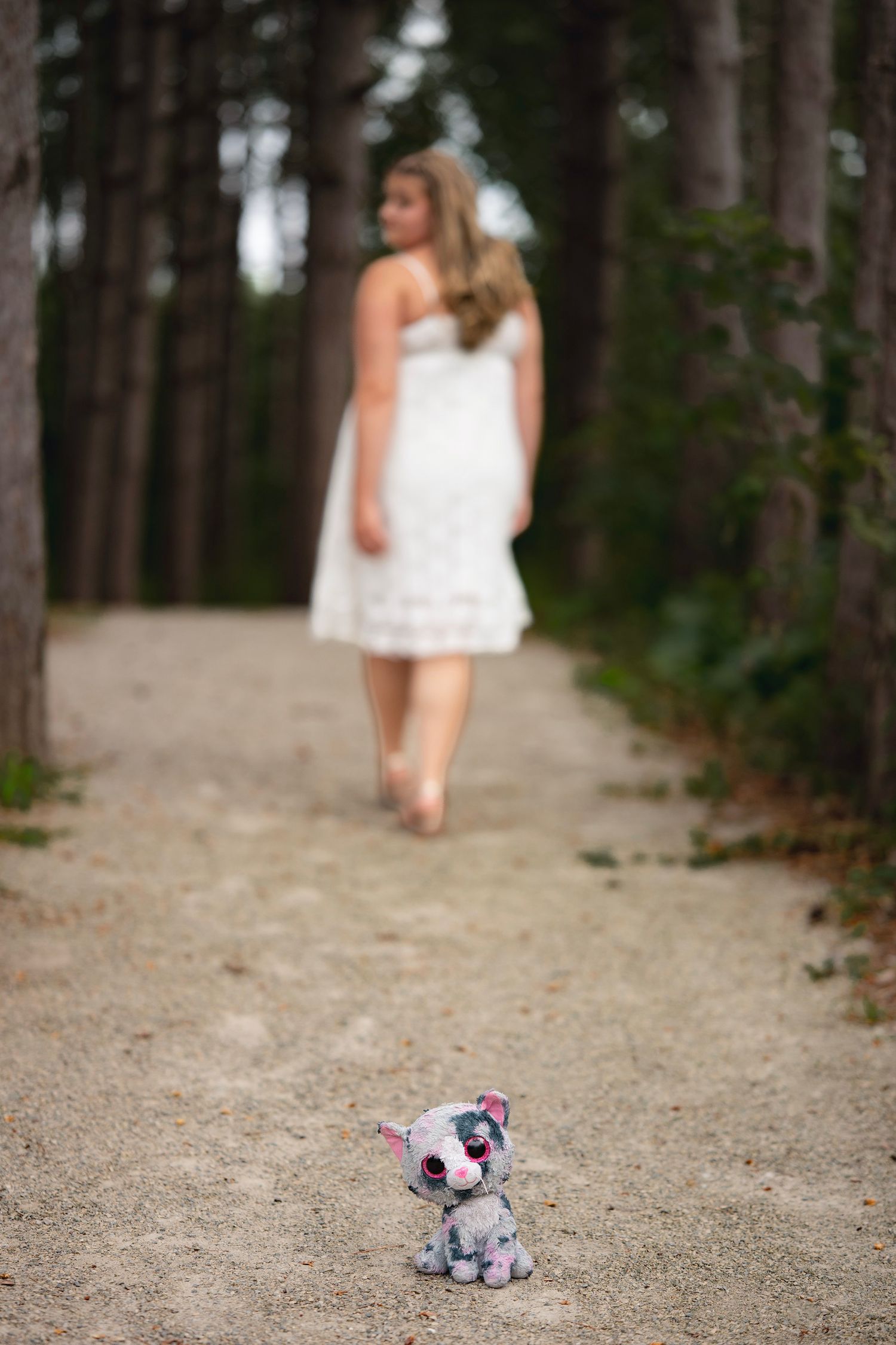 A small stuffed cat toy sits on a forest path as a woman in a white dress walks away among tall trees.