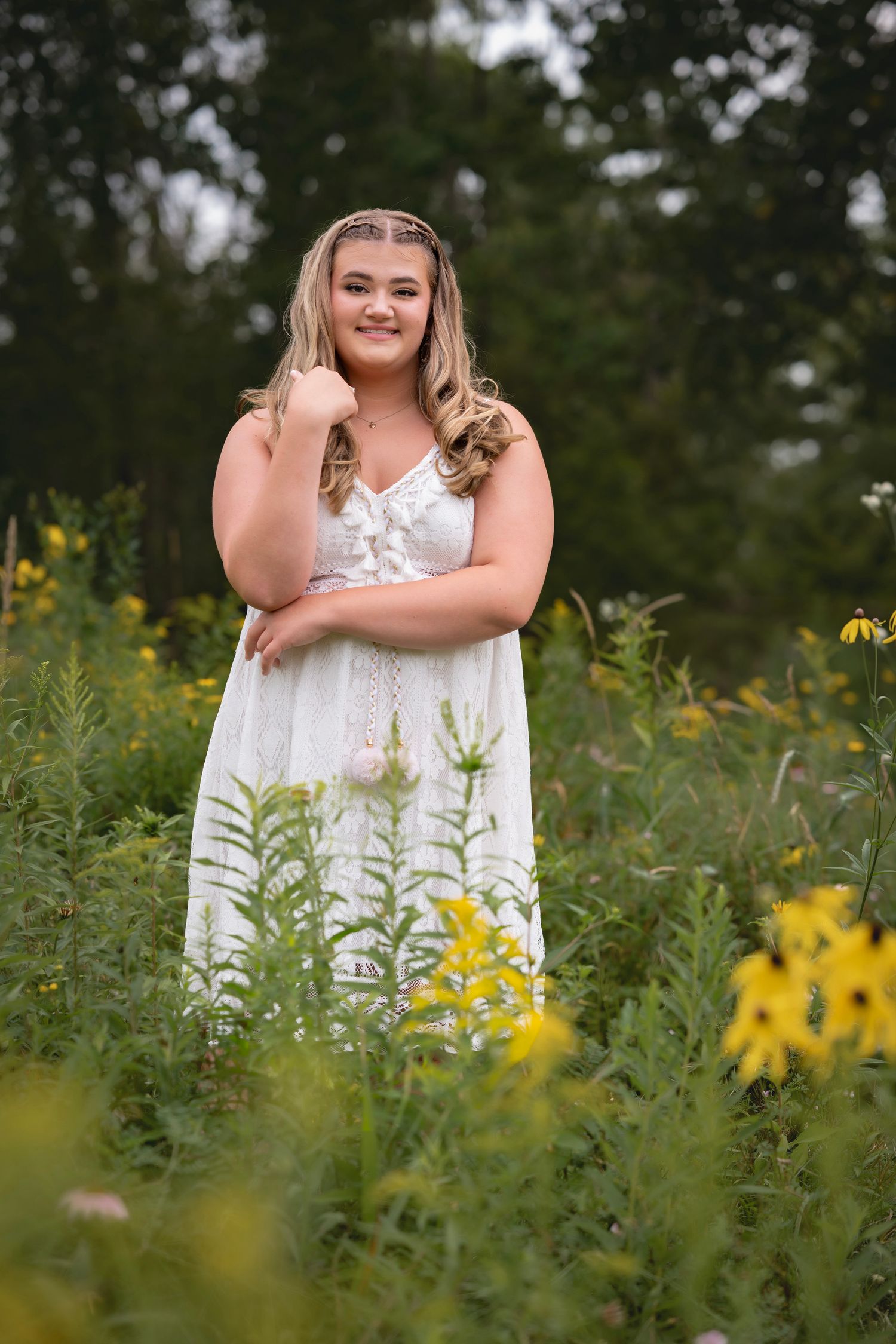 Young blonde woman in white sundress posing among yellow wildflowers in a lush green garden setting.