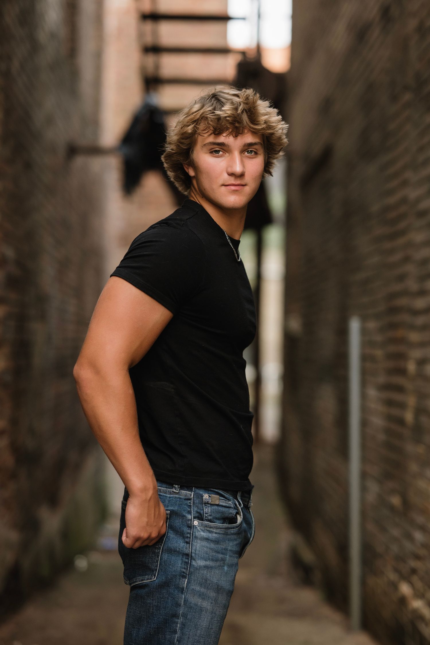 Person wearing black t-shirt and blue jeans poses in moody alleyway with brick walls.
