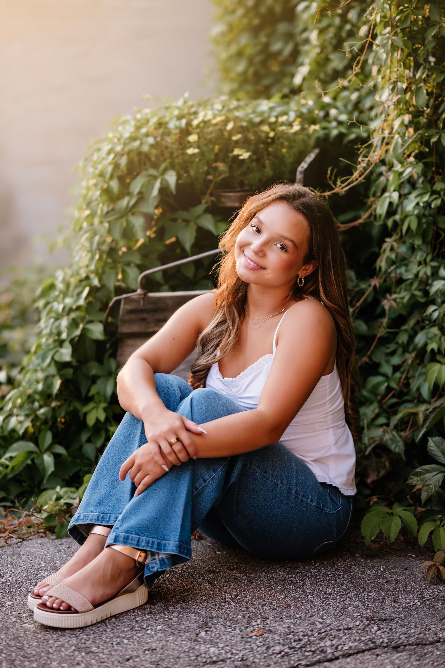 Young woman in white tank top and jeans sitting outdoors against lush green ivy wall in warm sunlight.