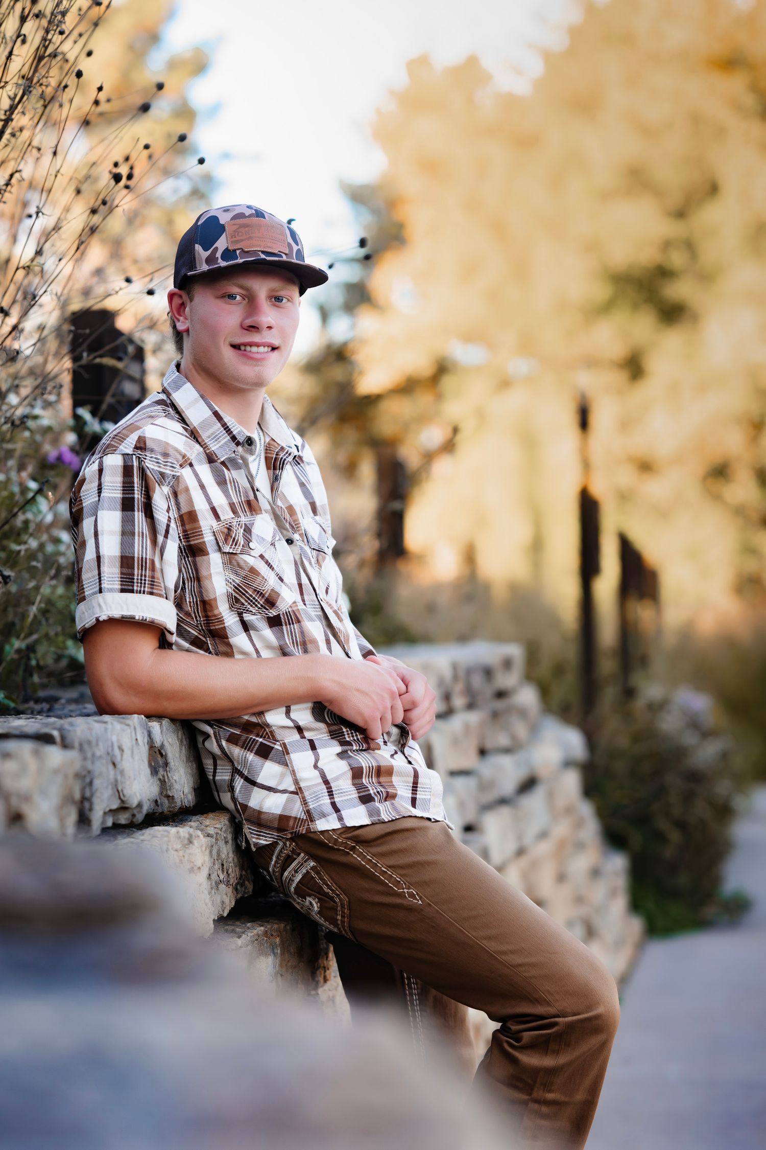 Person wearing plaid shirt and brown pants sits on stone wall in autumn outdoor setting.