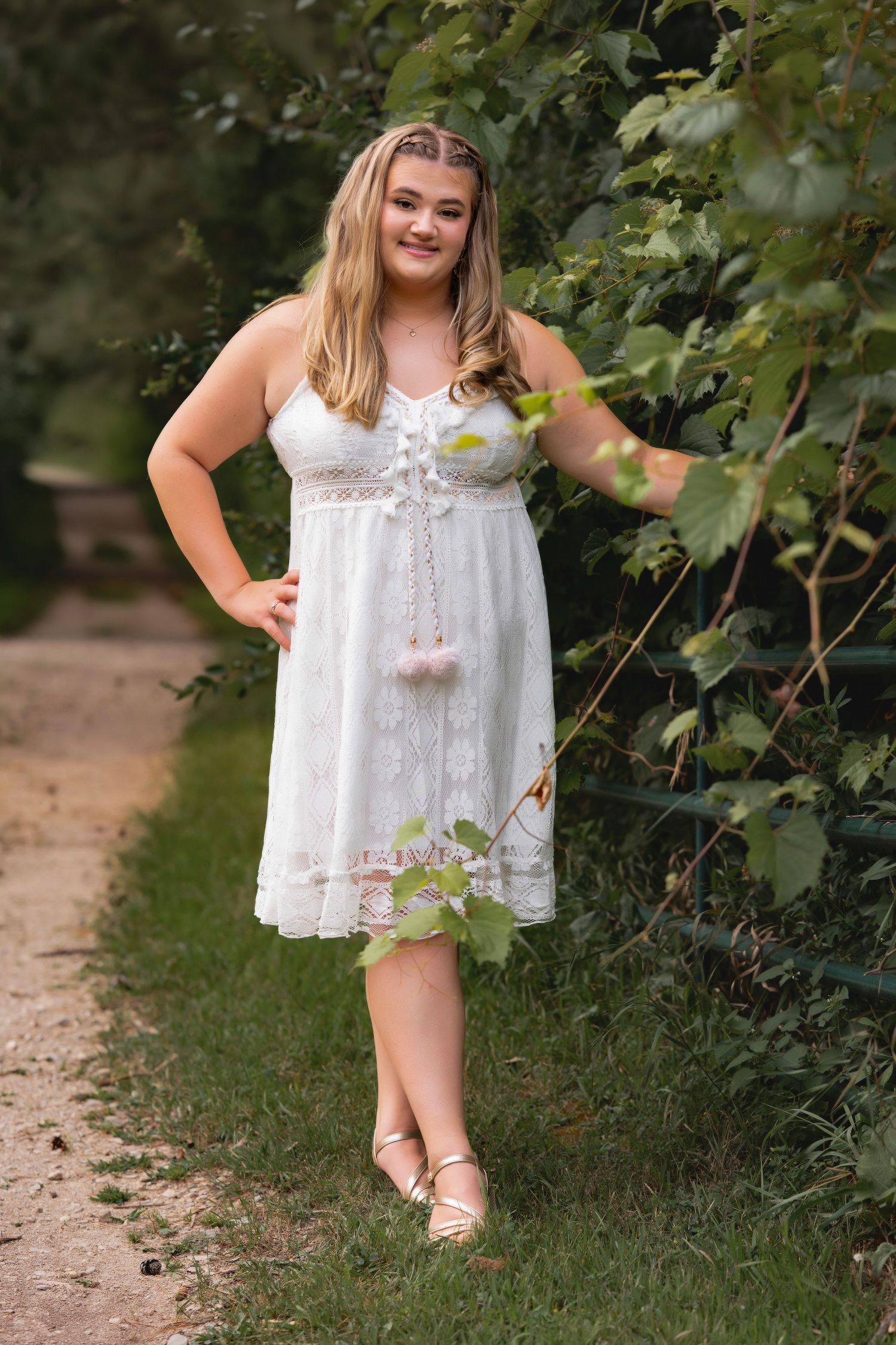Young woman in white lace dress poses outdoors near green foliage on a garden path.
