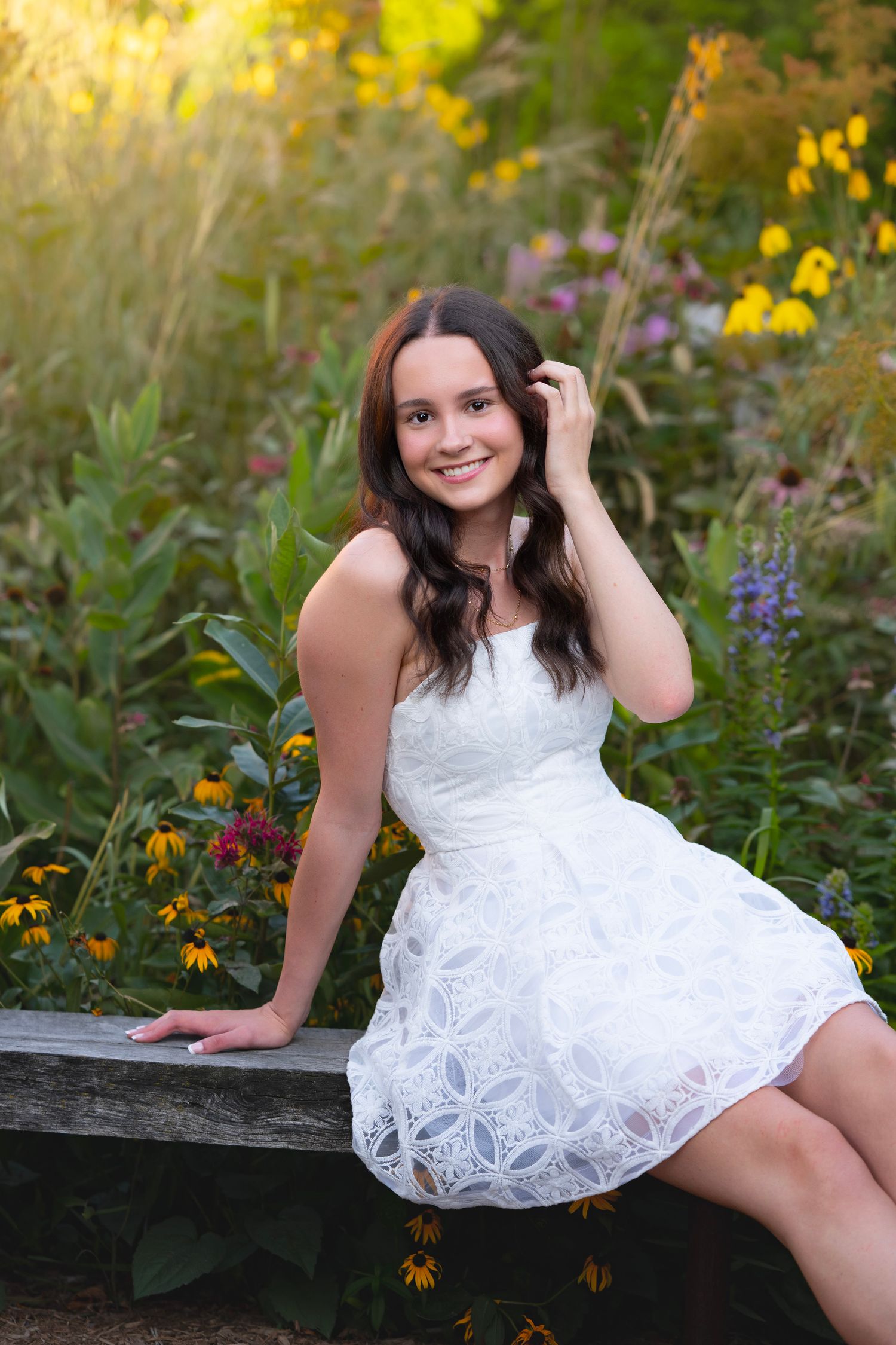 Young woman in white lace dress sitting on wooden bench surrounded by colorful wildflowers in golden hour light.