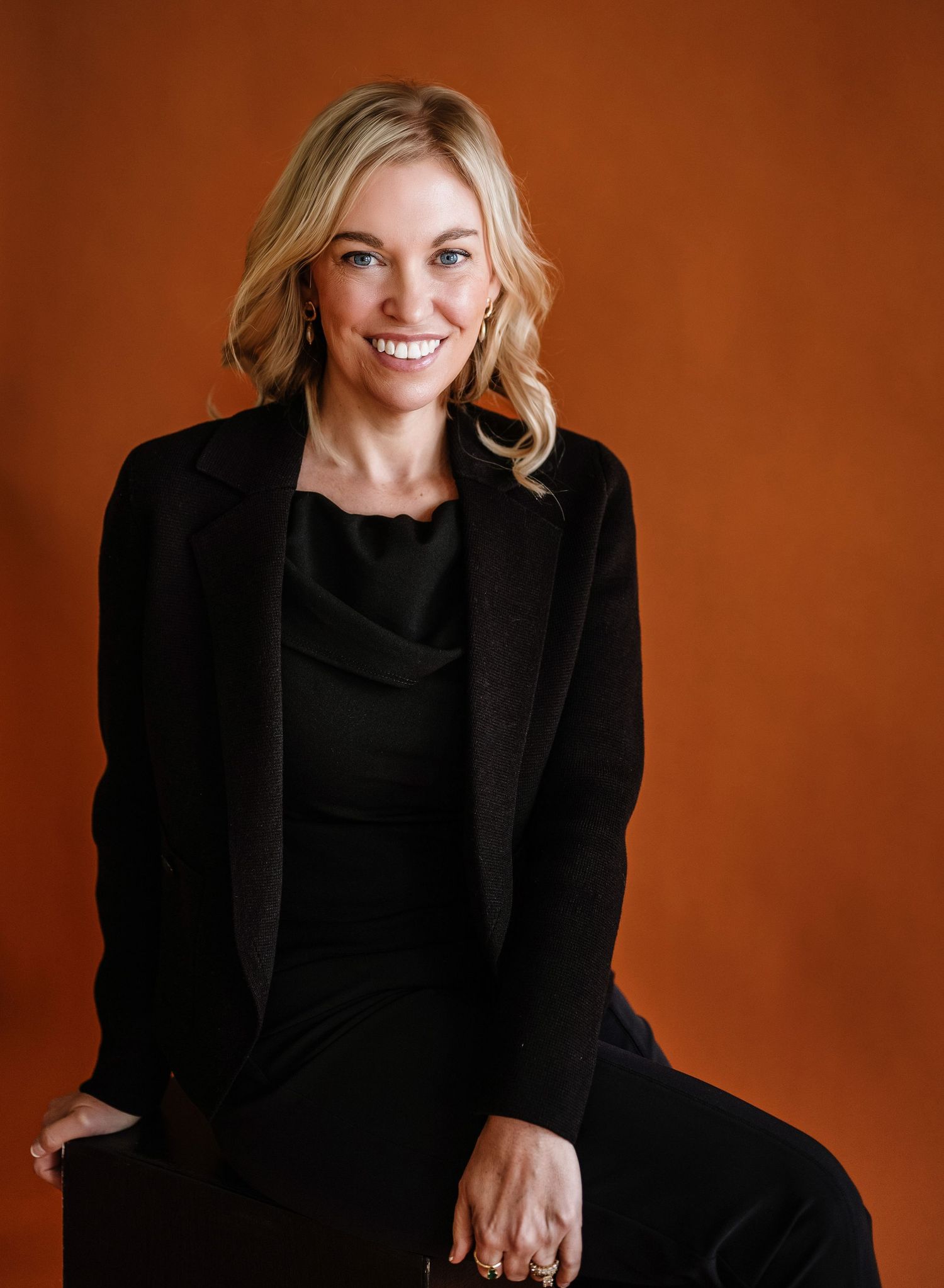 Professional studio portraits against an orange background showing someone in a black blazer with blonde hair posing expressively.