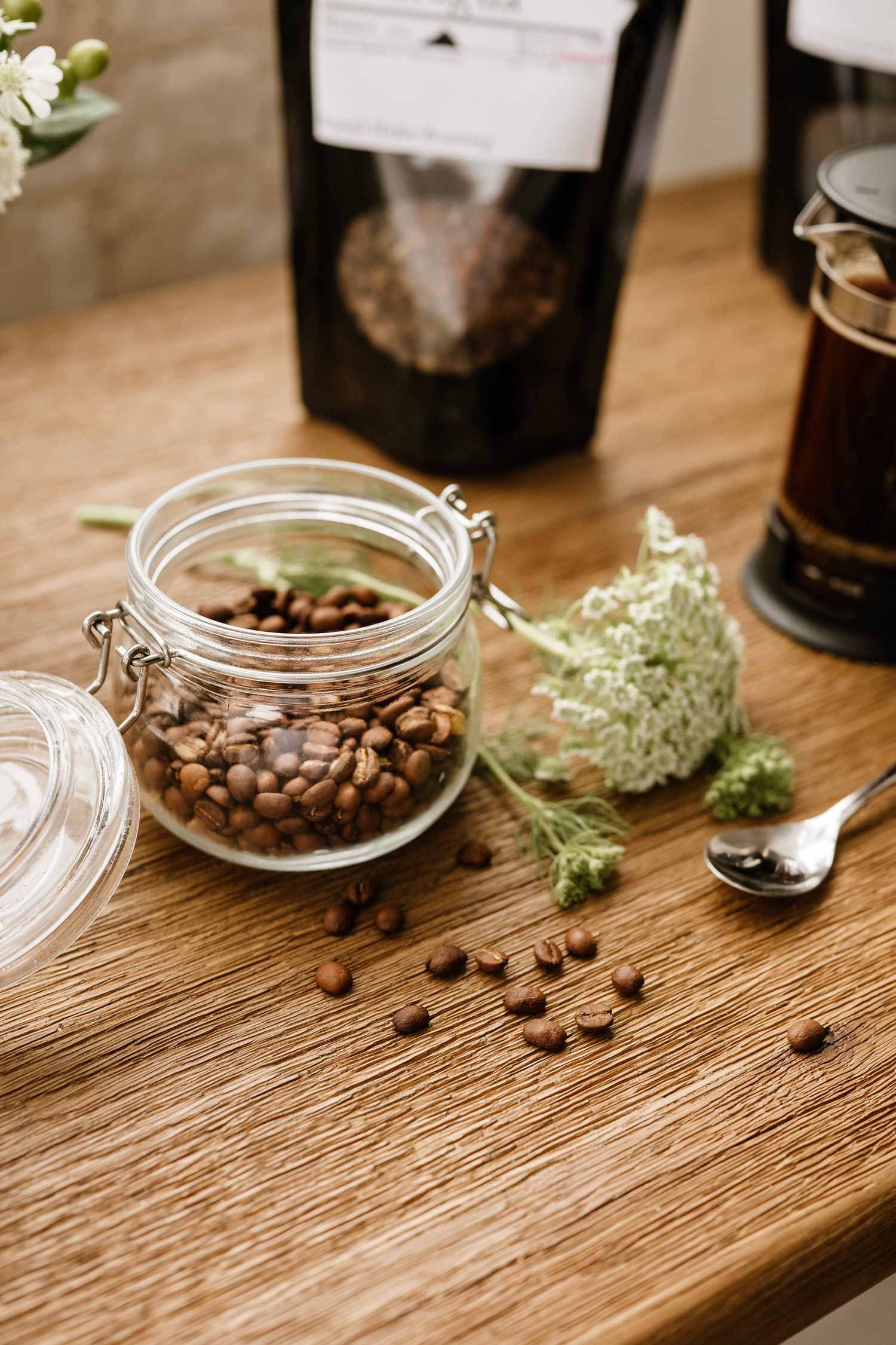 Glass jar filled with coffee beans on wooden table with white flowers, spoon, and coffee packaging in background.