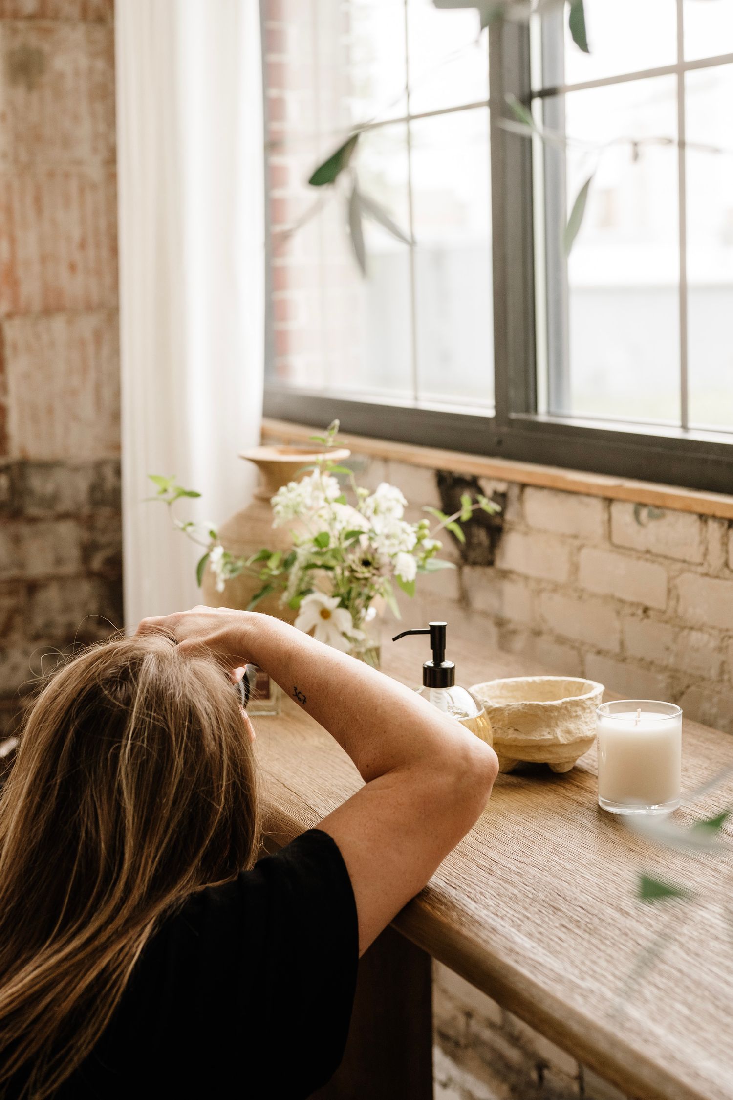 Woman with blonde hair relaxing at wooden vanity table with flowers, candle, and soap dispenser by window.