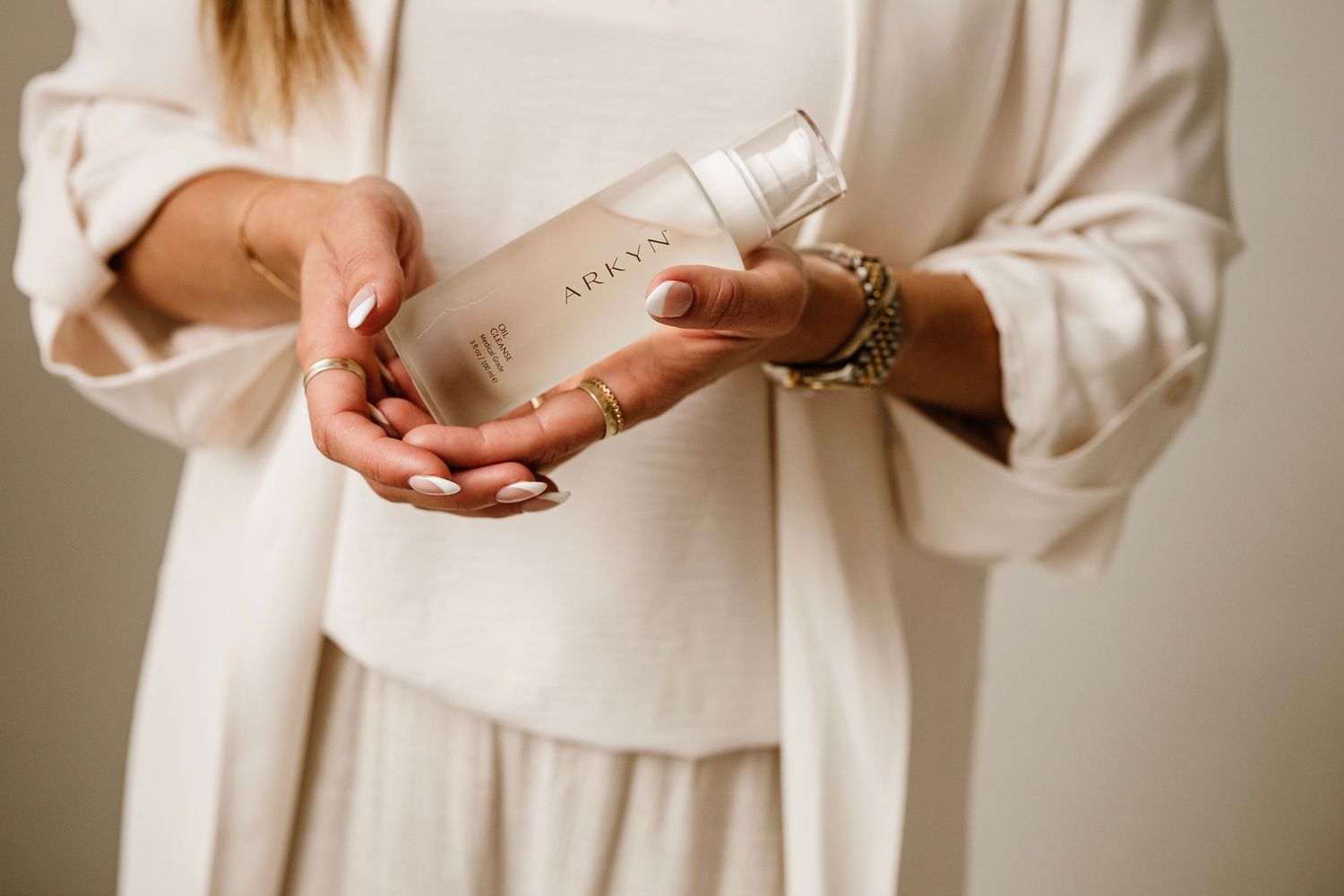 Woman in white outfit holding an ARKAN skincare bottle product with elegant gold jewelry.
