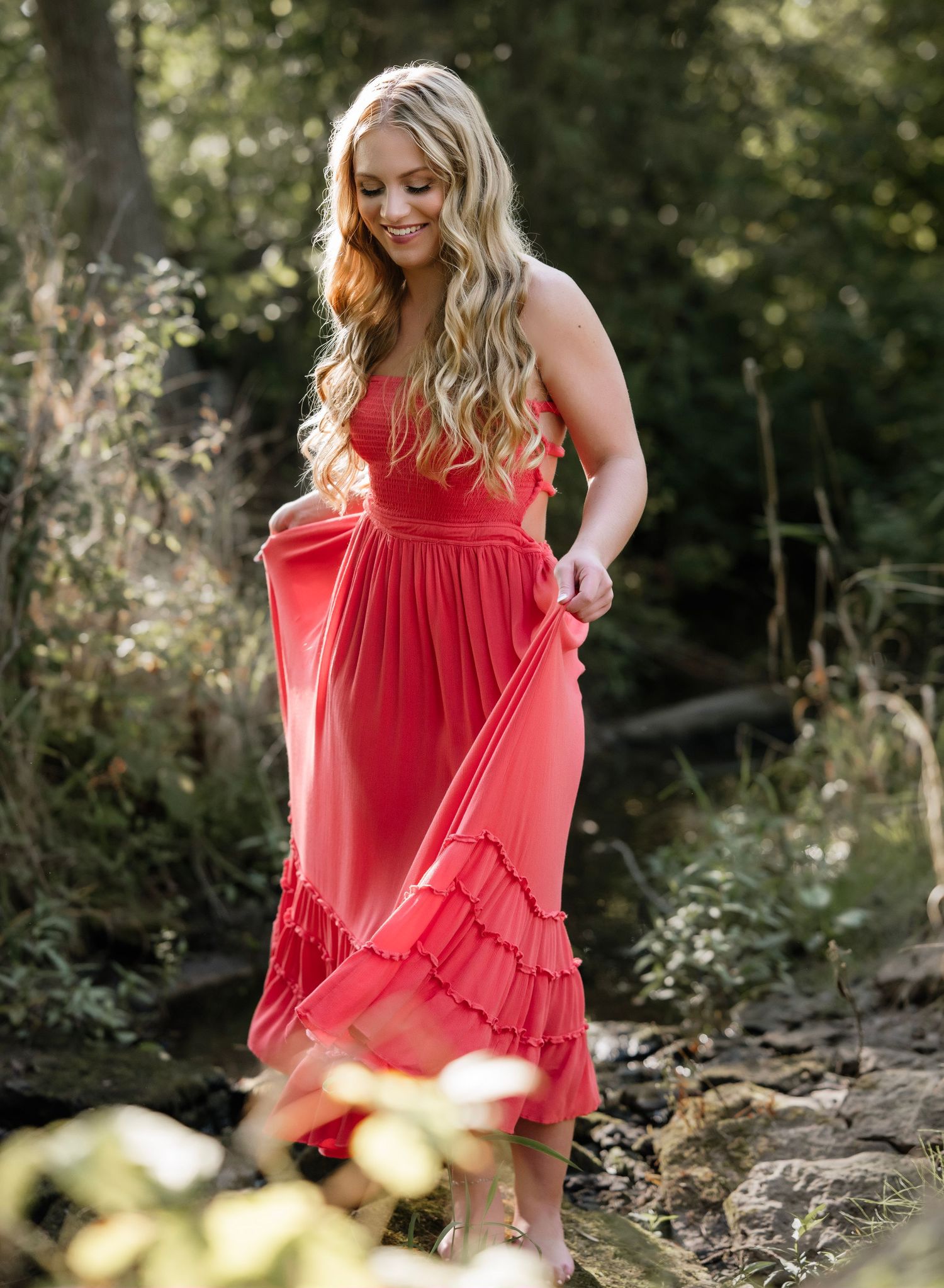 Figure in a flowing coral dress poses thoughtfully on natural stone formations in a sunlit forest setting.