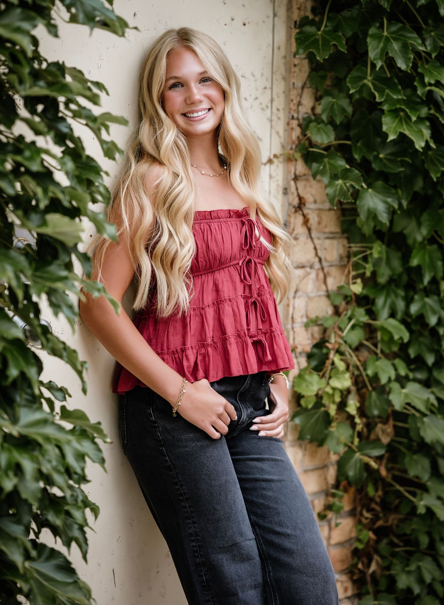 Smiling blonde woman in red ruffled top and black jeans posing against ivy-covered brick wall outdoors.