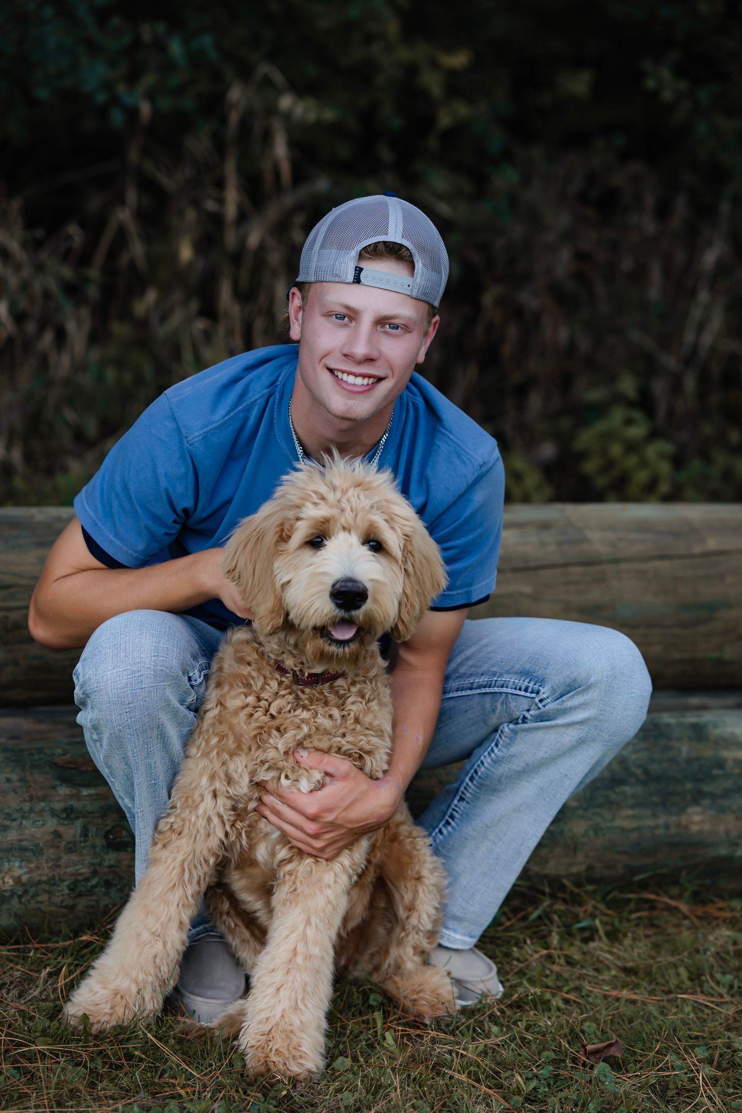 A young person in a blue shirt and baseball cap crouches next to their happy goldendoodle dog on grass.