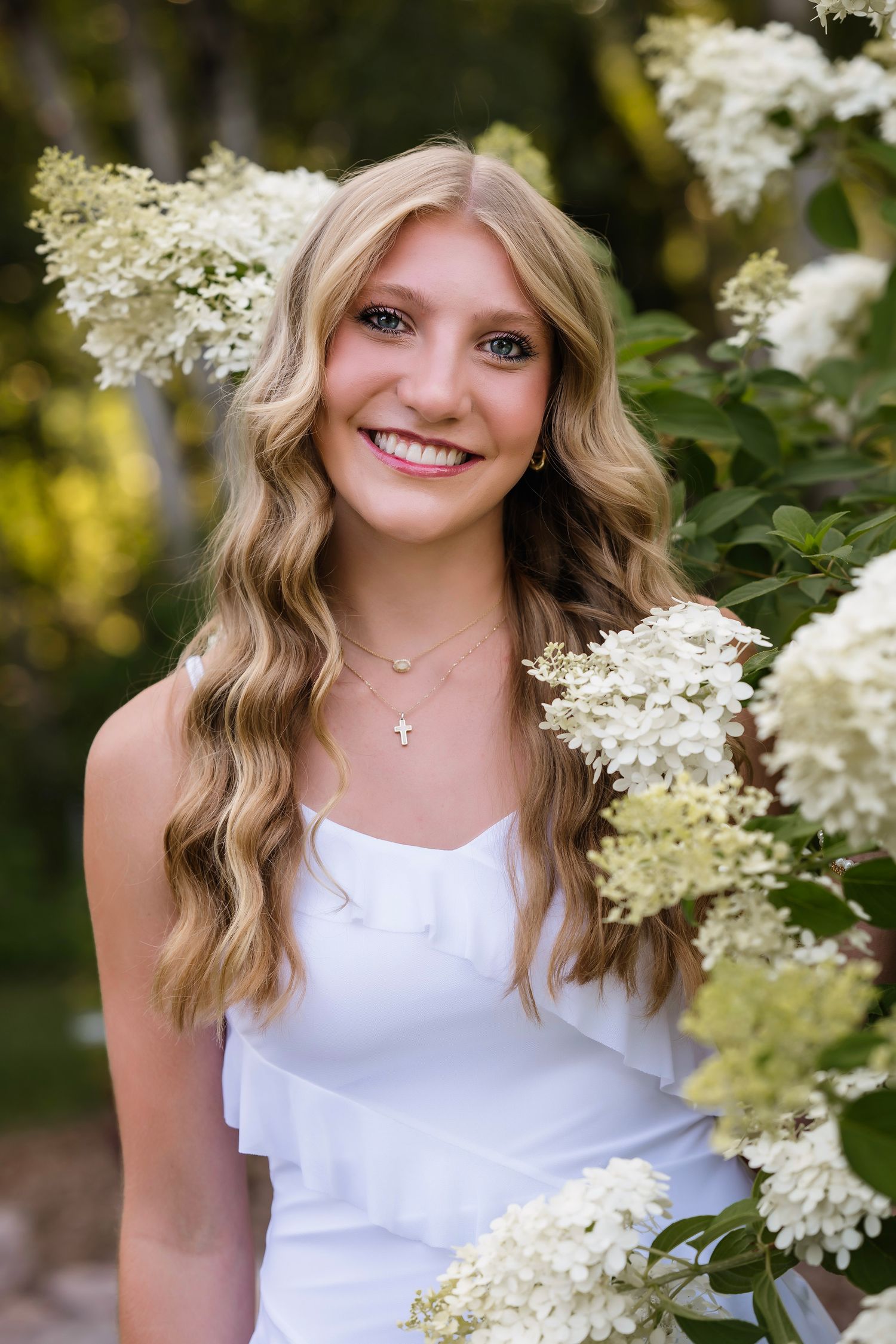 Young blonde woman in white dress smiling among white hydrangea blooms, wearing a delicate cross necklace outdoors.