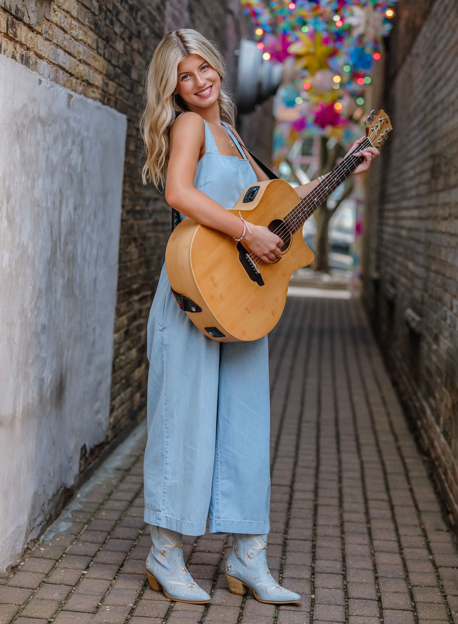 A musician in a light blue dress poses with an acoustic guitar against a brick wall with colorful balloons overhead.