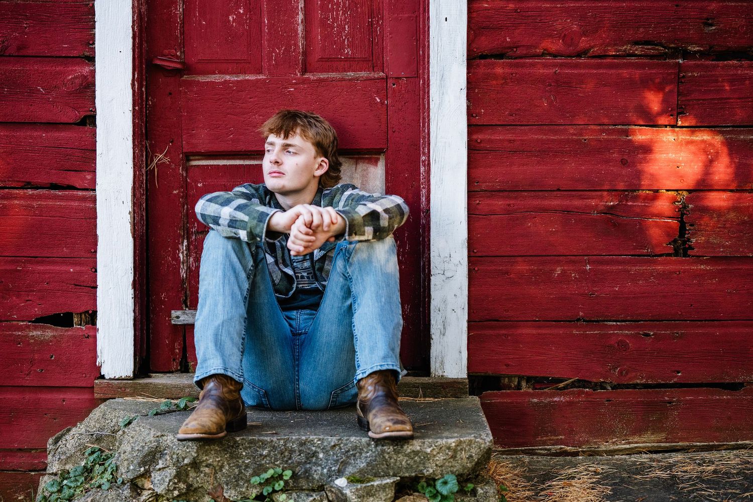 A person sits on a wooden porch with a red barn wall backdrop in rustic country setting.