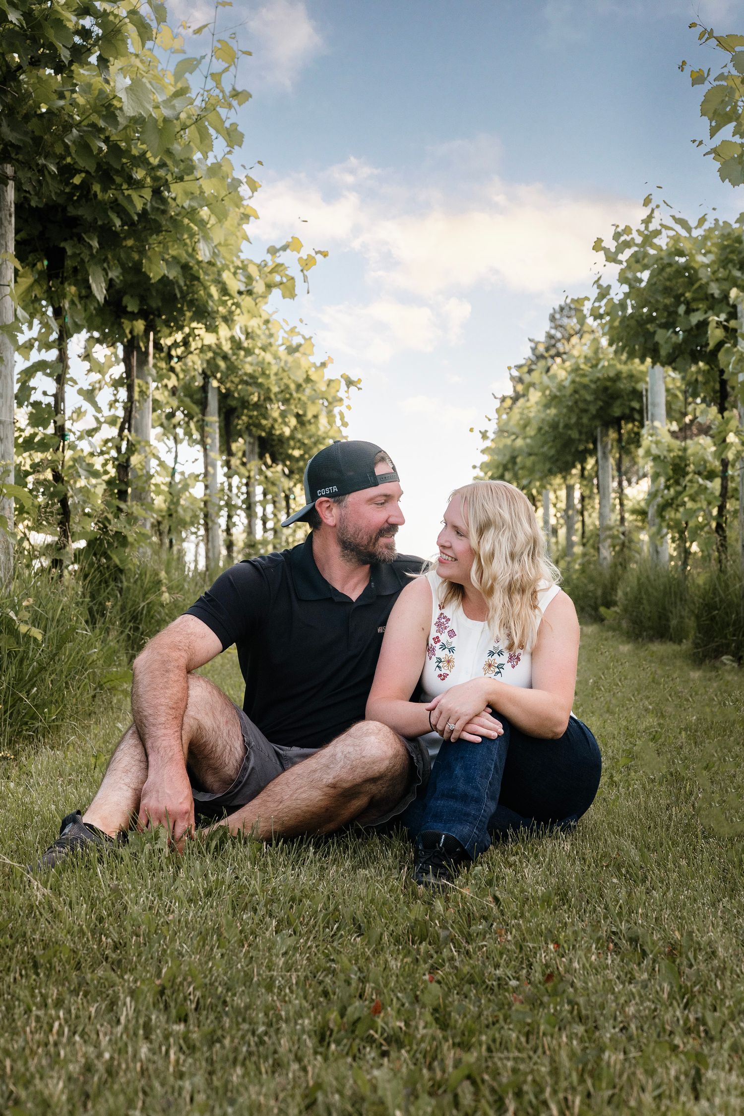 Couple sitting together on grass between vineyard rows, smiling at each other on a sunny day.