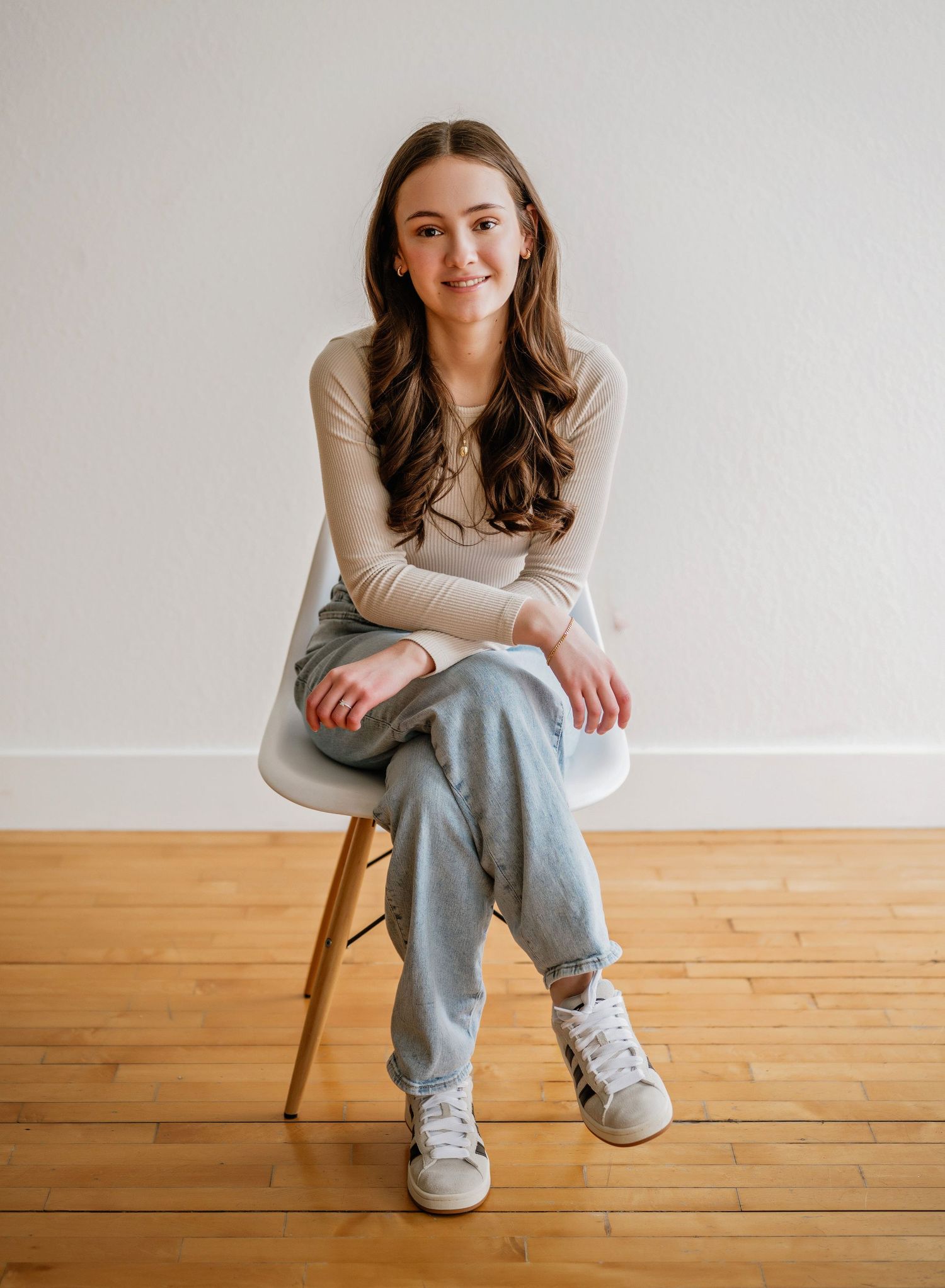 A person in a beige sweater and gray sweatpants sits casually on a wooden stool against a minimalist white background.