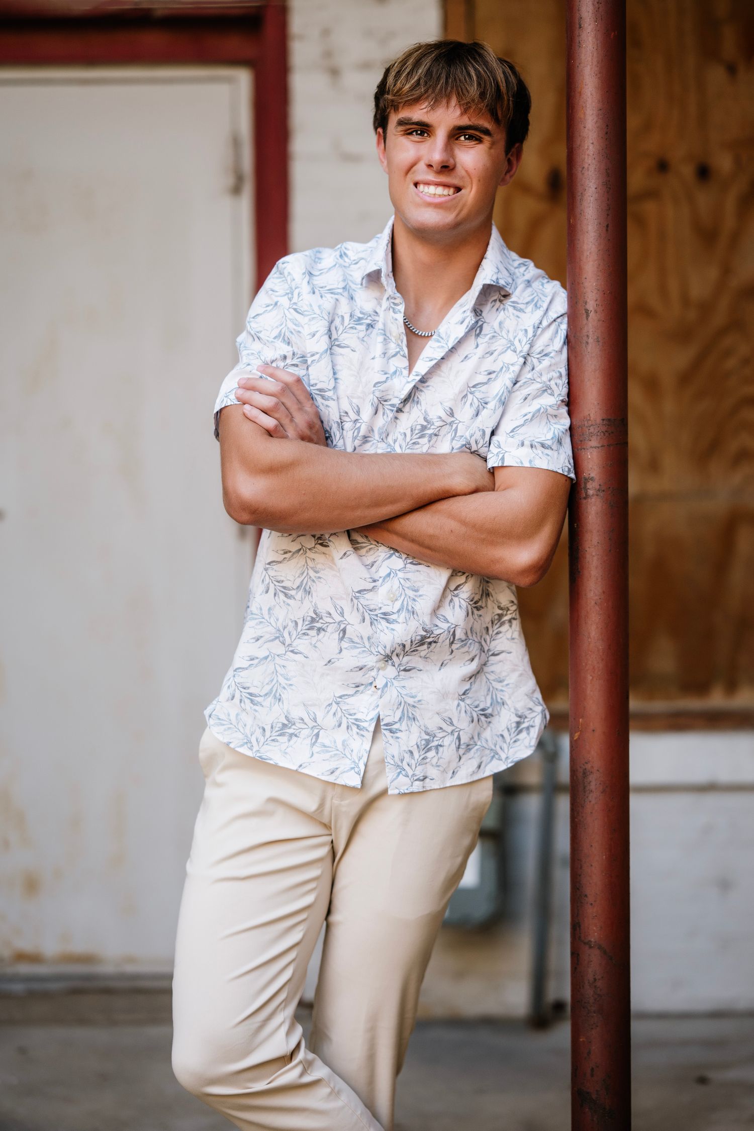 Person in white patterned shirt and khaki pants poses casually against rustic wooden backdrop in outdoor setting.