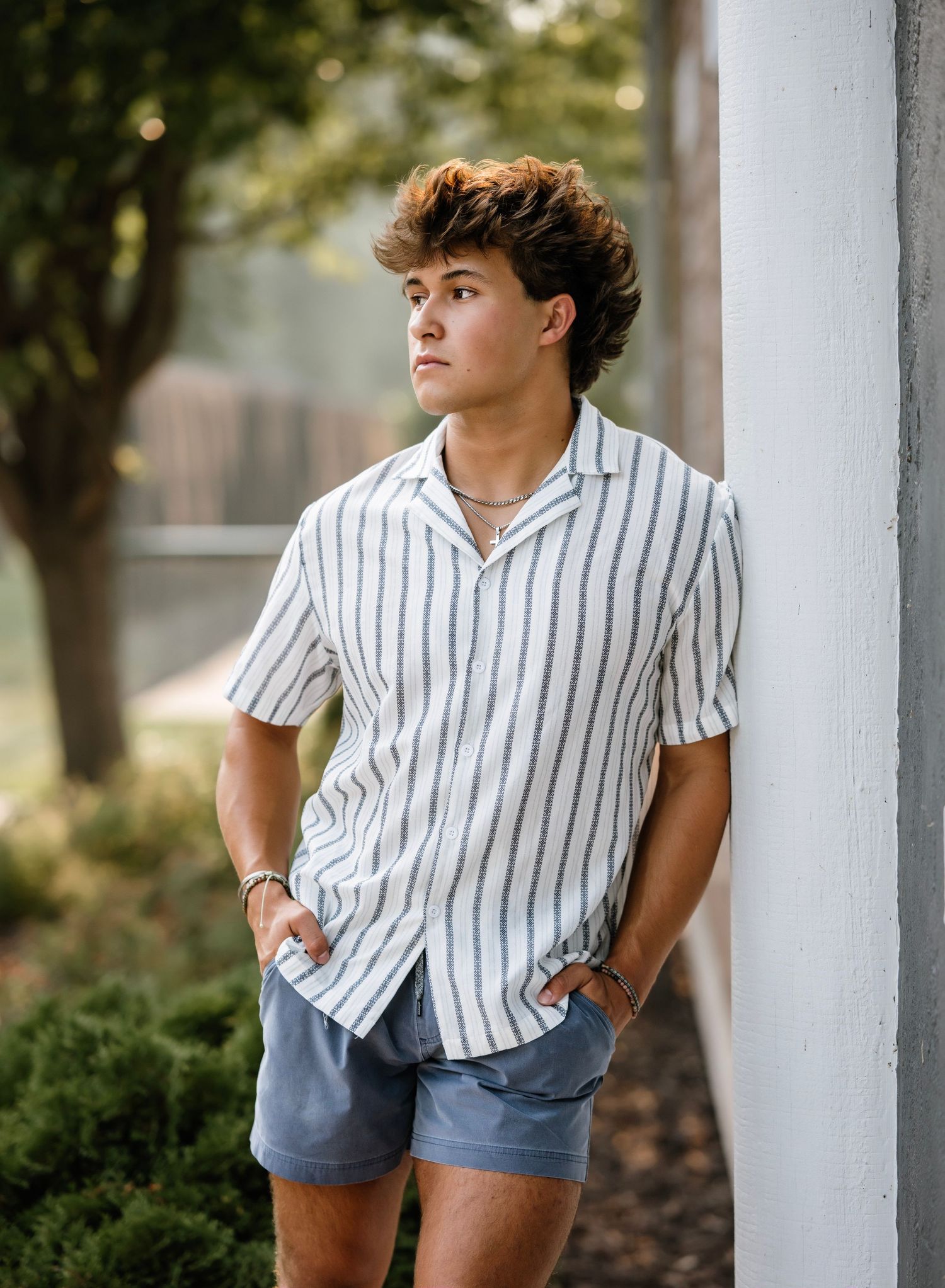 A person in a striped button-down shirt and denim shorts poses casually against an outdoor pillar in natural lighting.