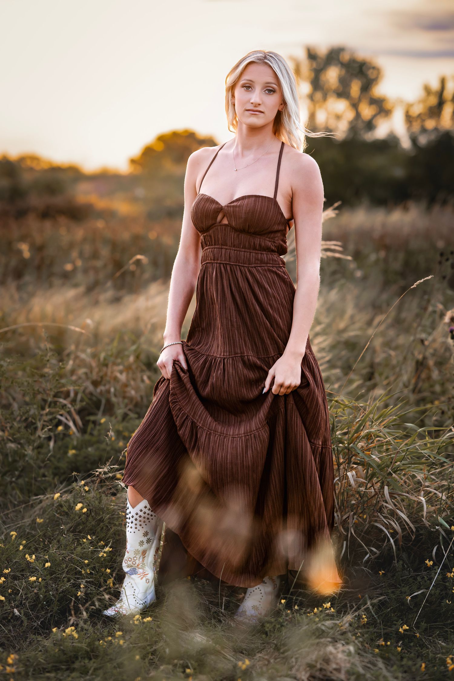 A person in a brown maxi dress and pink top poses in a wildflower field at sunset wearing cowboy boots.