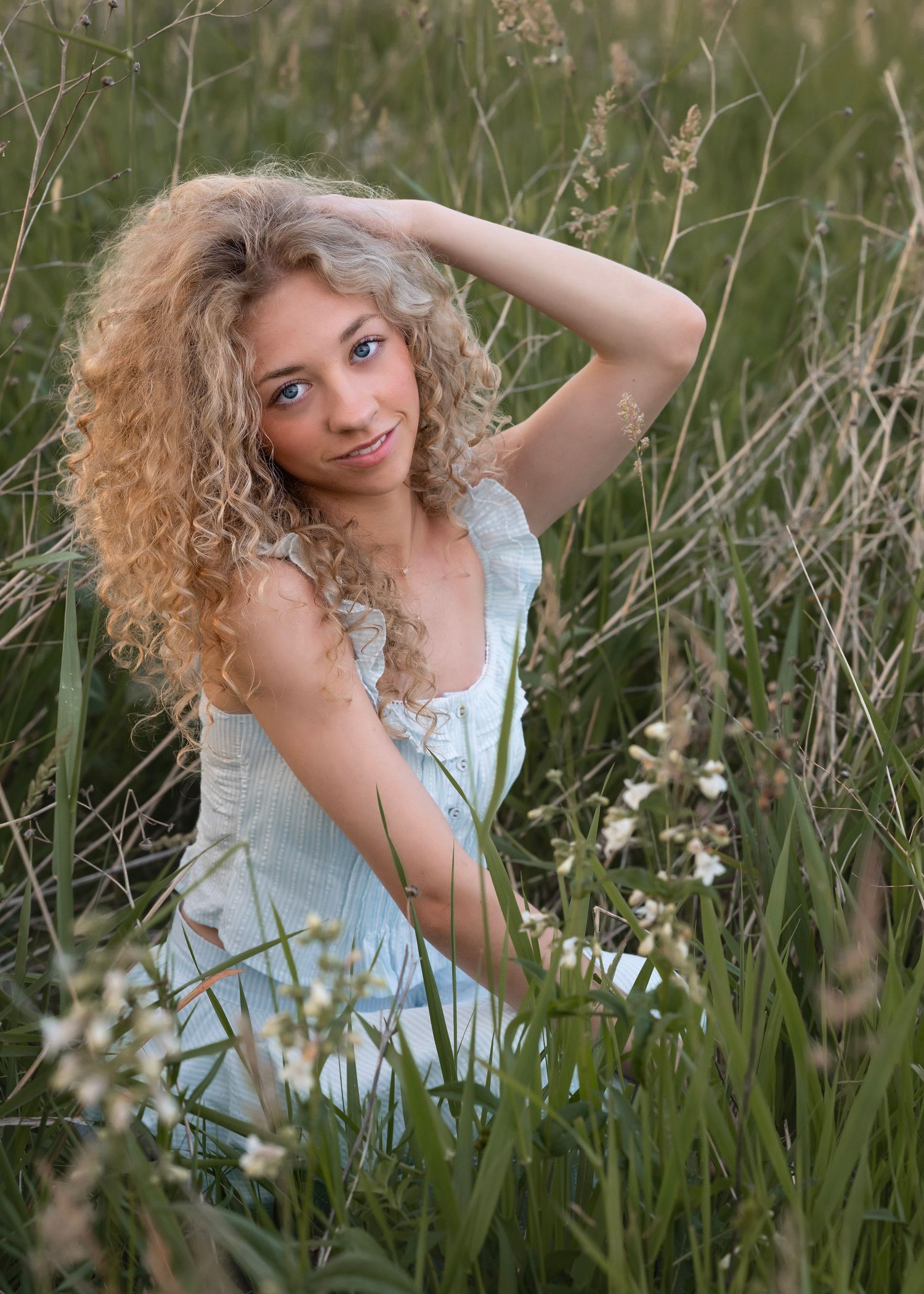 Young woman with curly blonde hair sitting in a field of tall grass and wildflowers wearing a light blue dress.