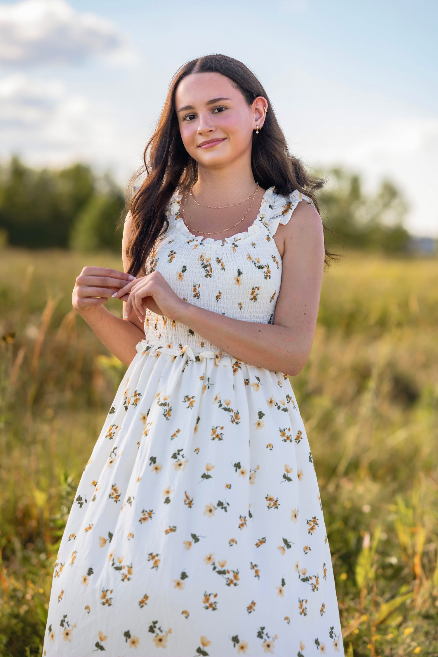 Young woman in white floral sundress standing in a golden field during sunset, smiling softly at the camera.