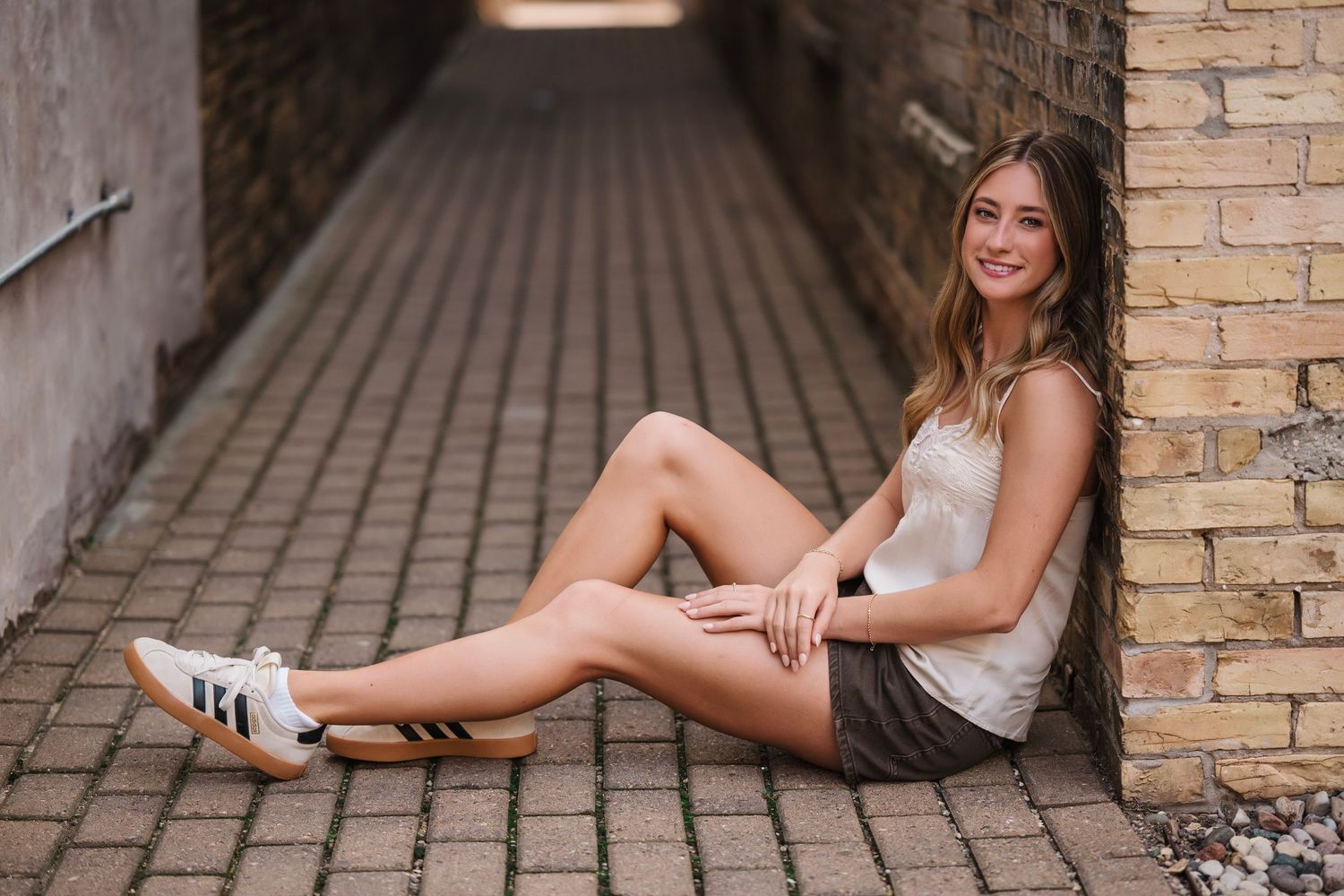 Young woman in white top and shorts sitting against brick wall in alley, smiling, wearing Adidas sneakers.